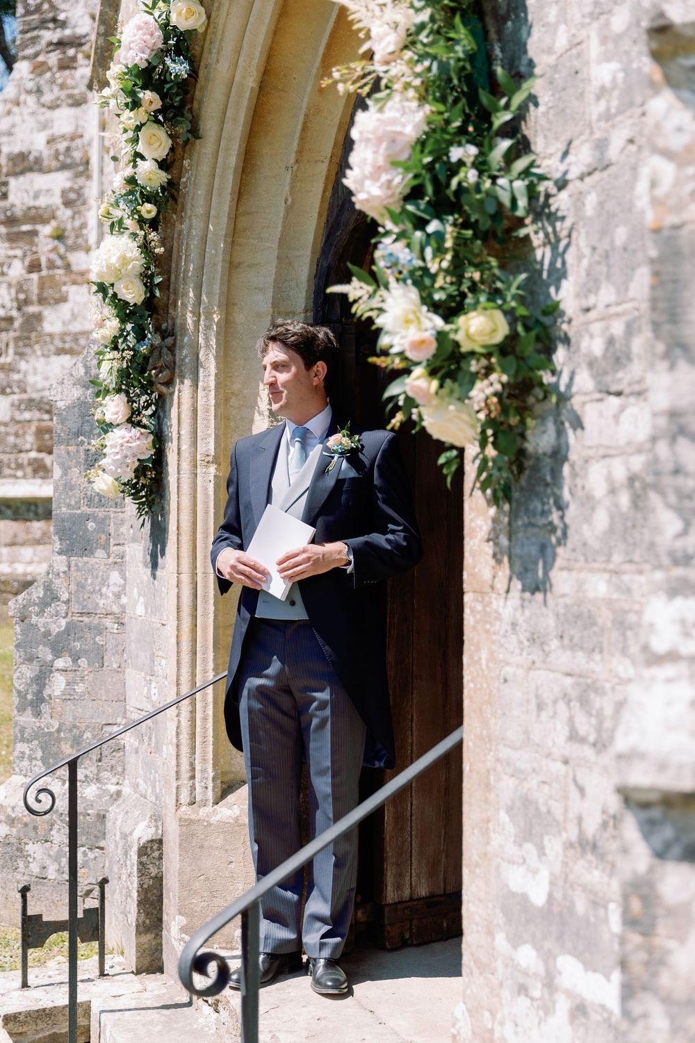 A groom waits in formal attire at a stone church entrance decorated with white floral arrangements.