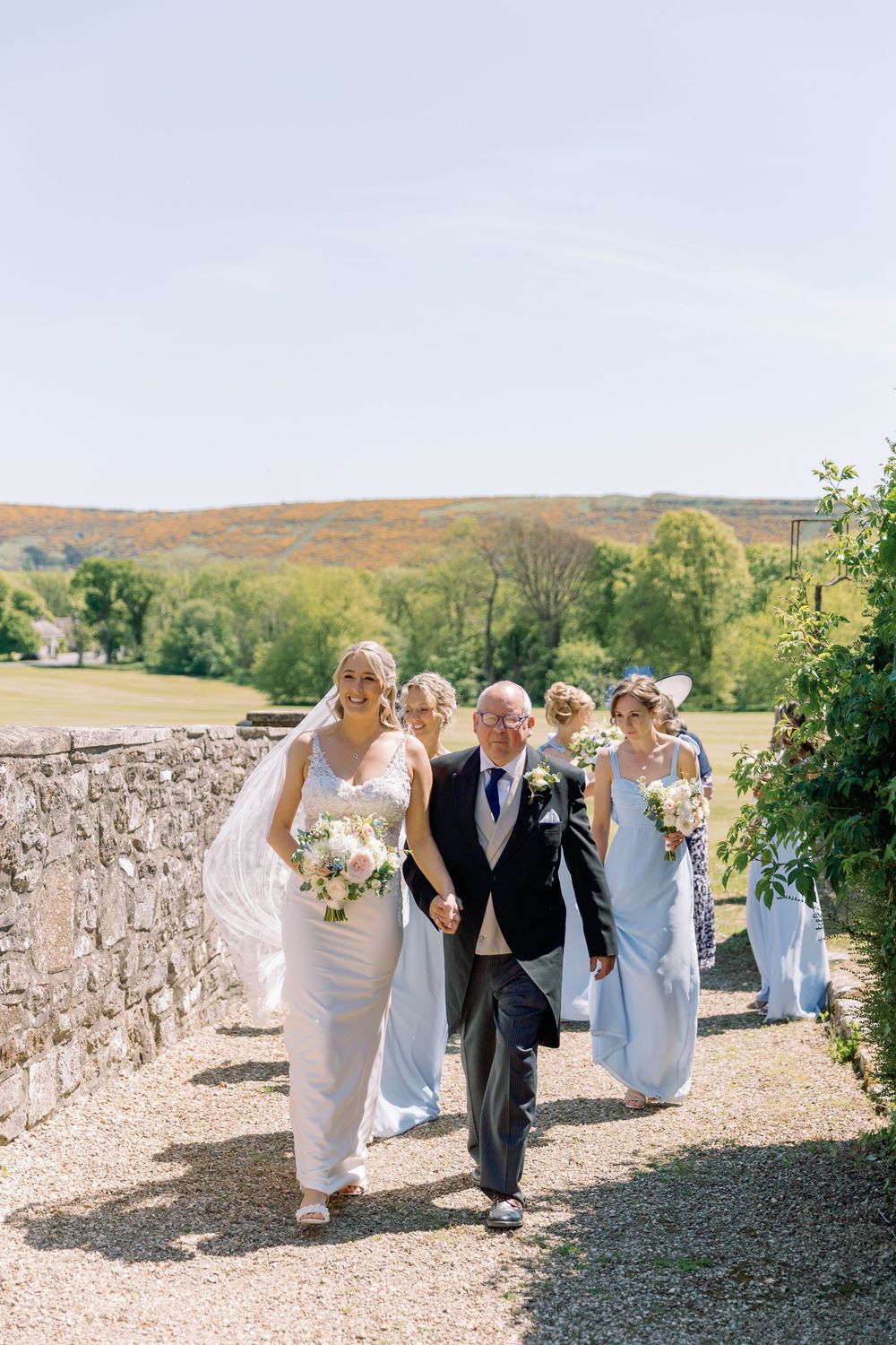 Wedding party walks along a stone wall path at an outdoor venue with rolling hills in the background.