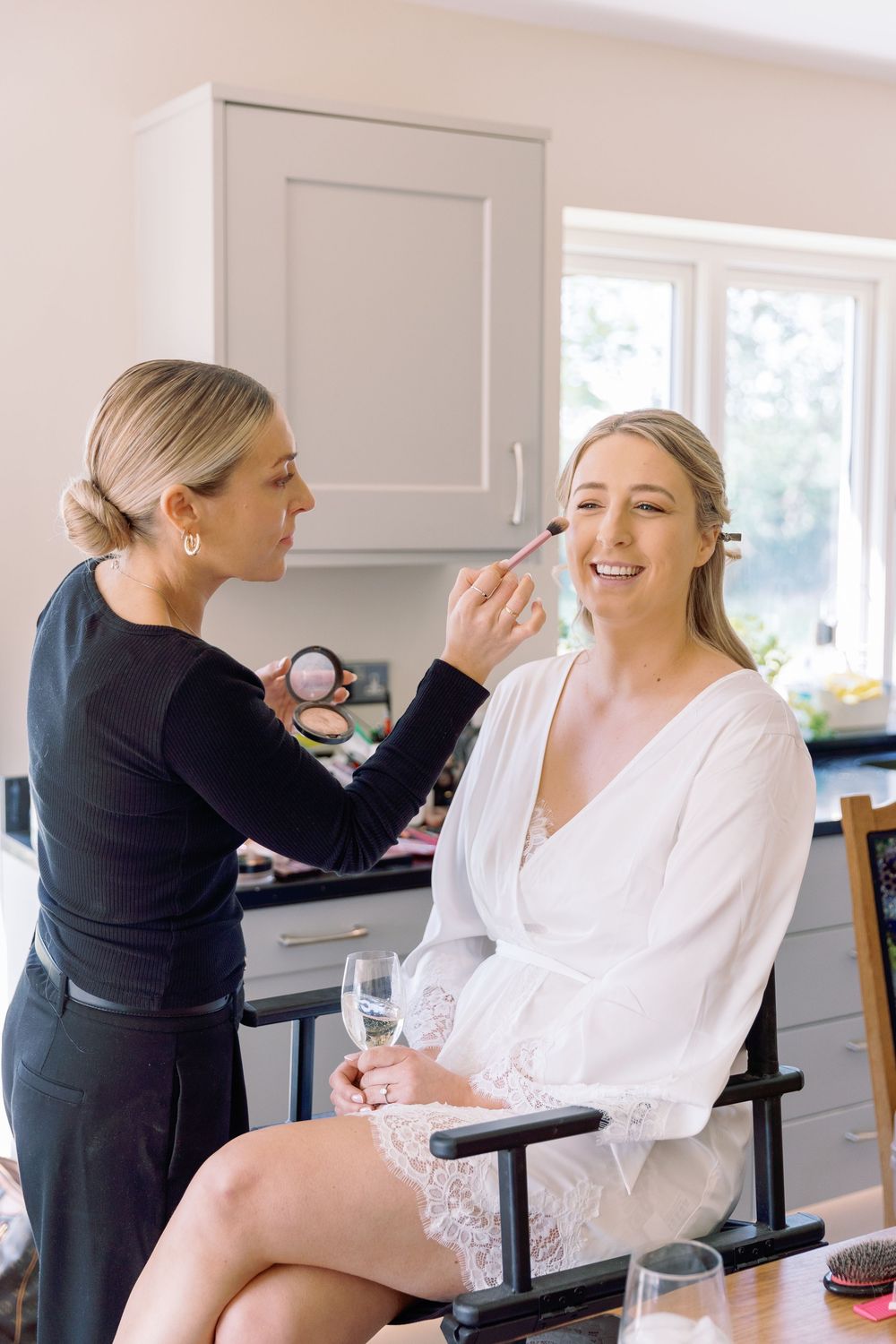 Makeup artist in black applying cosmetics to client in white robe during wedding preparation session.