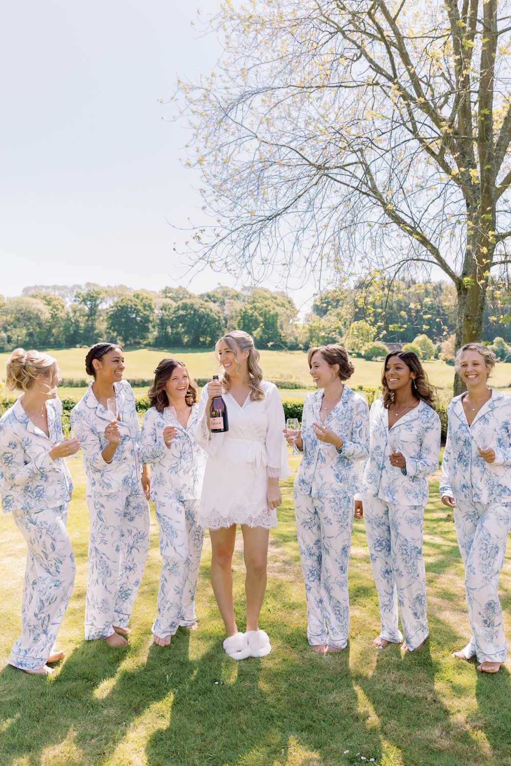 Wedding party wearing matching floral robes poses playfully on a sunny lawn with trees in the background.