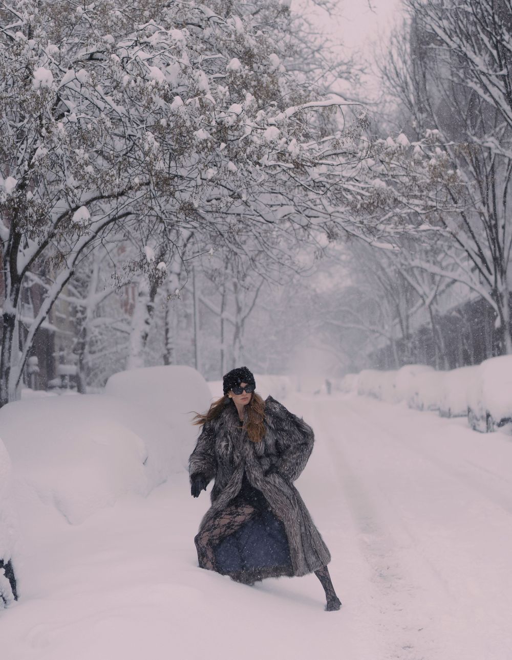 A person in winter clothing jumps playfully in deep snow on a tree-lined path during a snowy winter day.