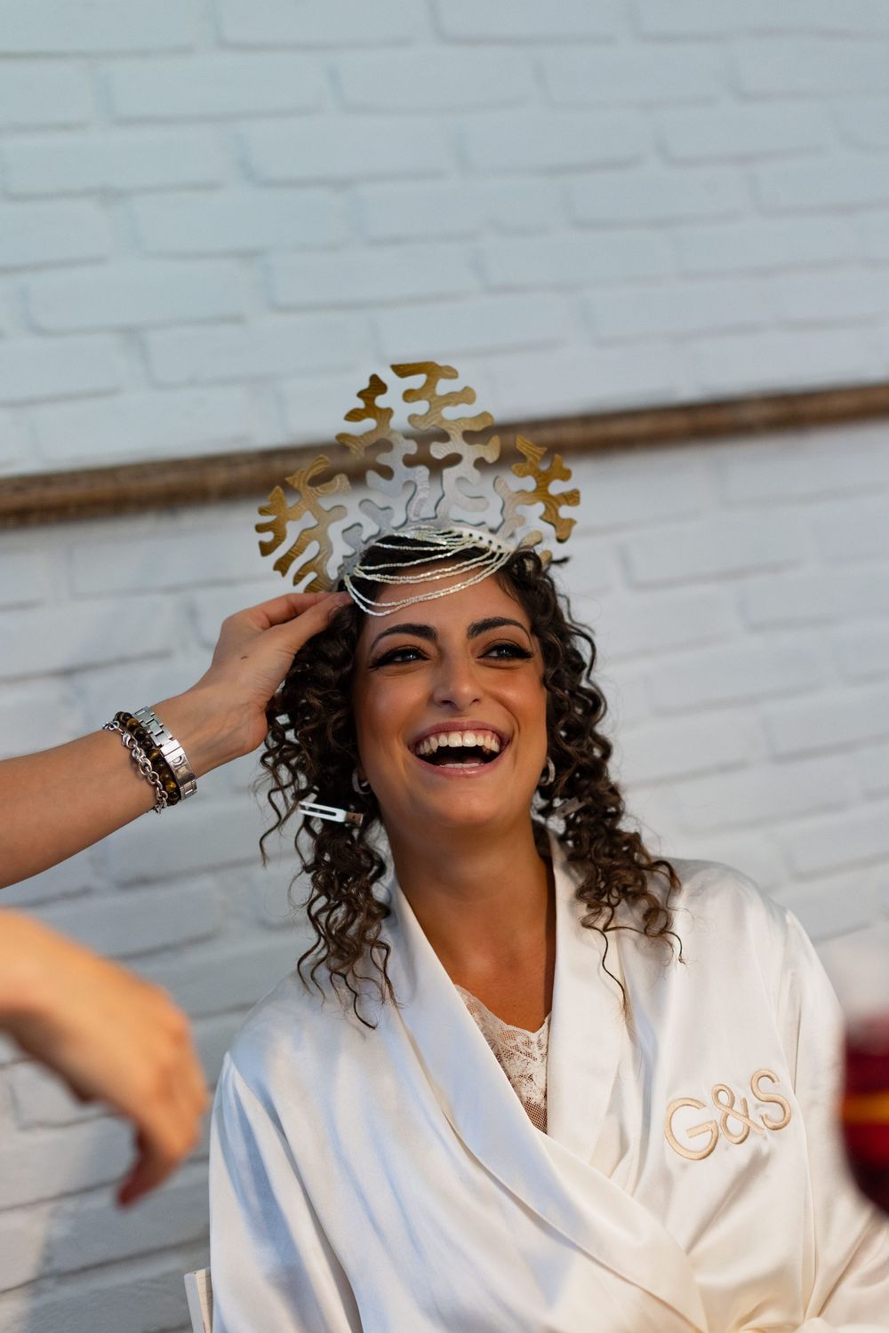A joyful moment captured during wedding preparations as a woman in a white robe laughs while wearing a decorative headpiece.