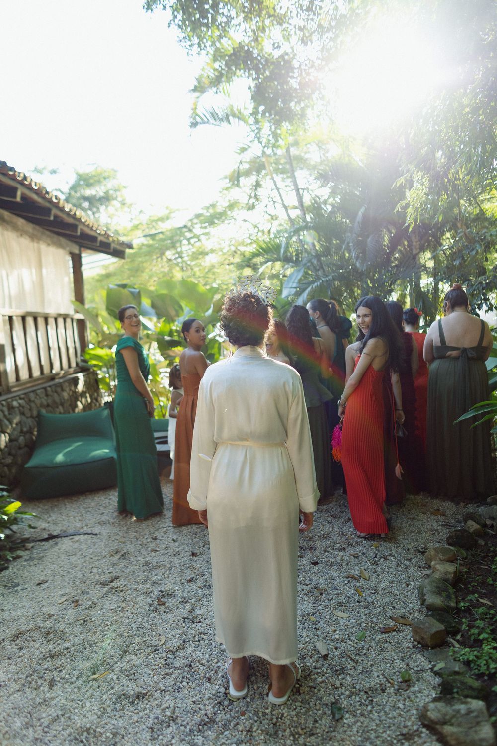 Wedding guests in colorful formal attire gather on a gravel path surrounded by tropical gardens and trees.