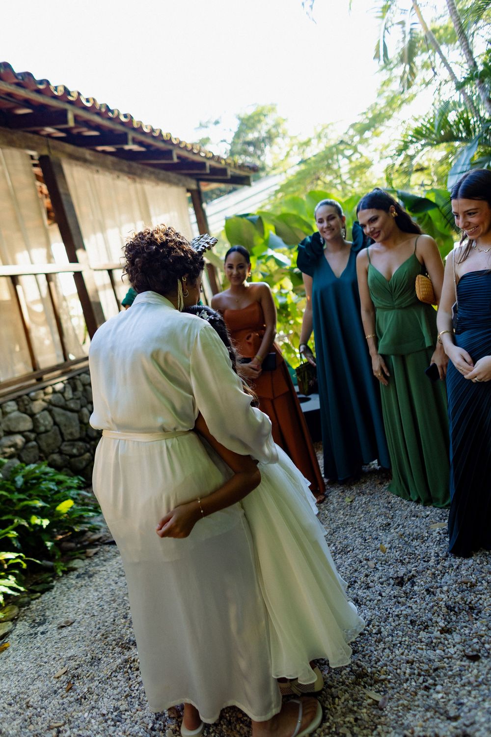 A couple in white attire dances together at an outdoor wedding reception on a gravel path surrounded by greenery.