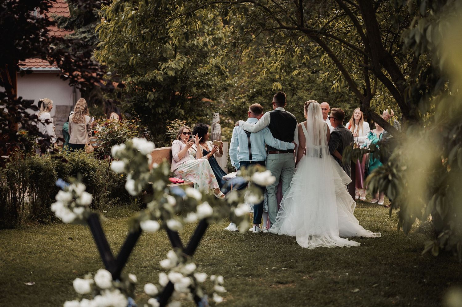 An outdoor garden wedding ceremony with white floral decorations and guests gathered on a grassy lawn.