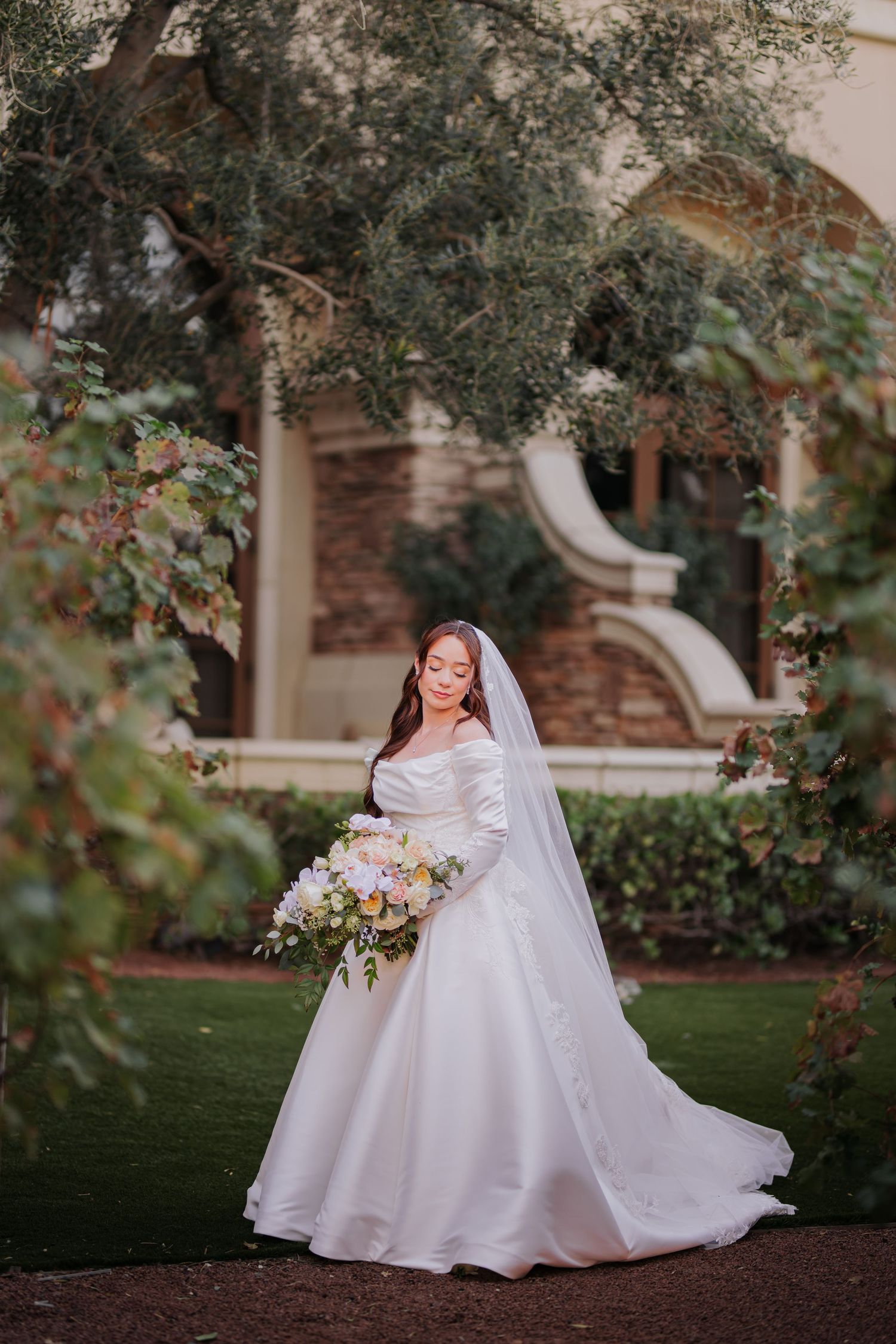 Bride in a white ball gown holding a floral bouquet, standing in a lush garden with elegant stone architecture at Green Valley Ranch.
