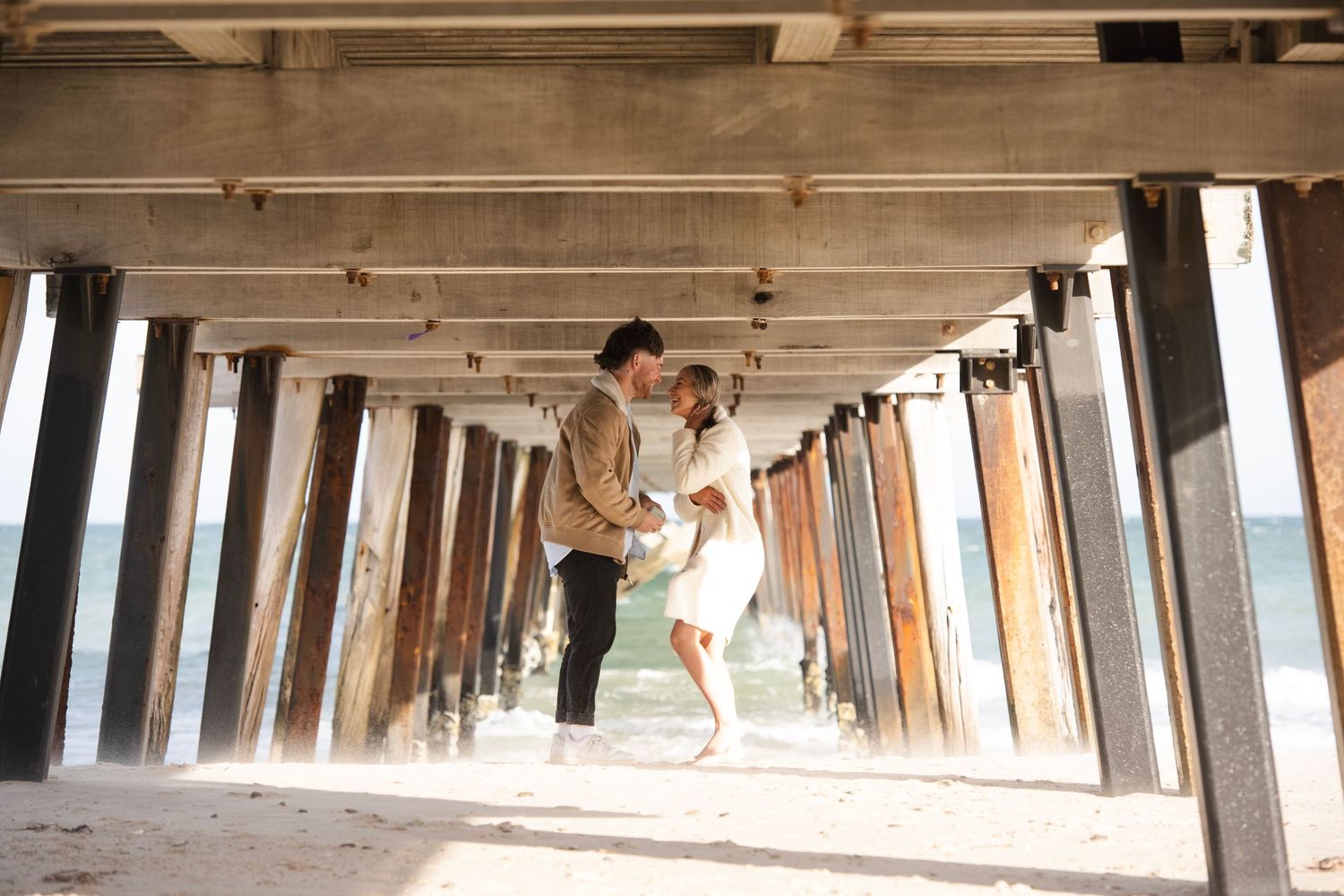 Couple sharing a romantic dip kiss under a beach pier with ocean waves in the background.