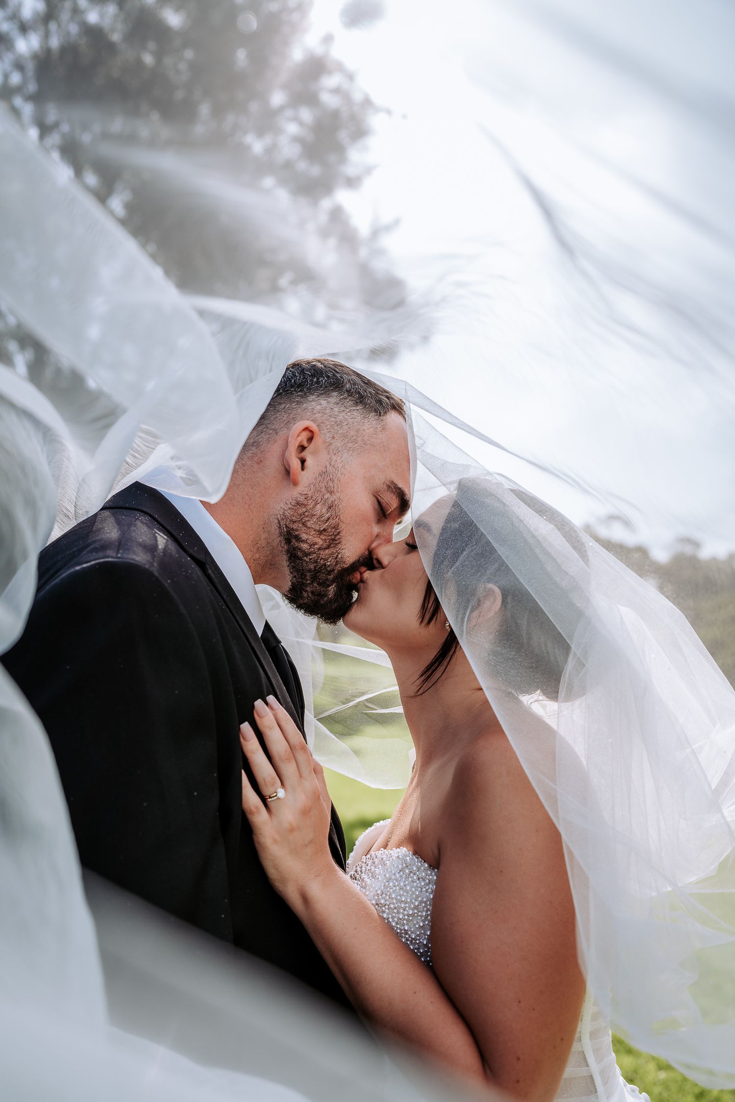 Romantic wedding couple sharing intimate moments under a flowing veil in an outdoor setting.