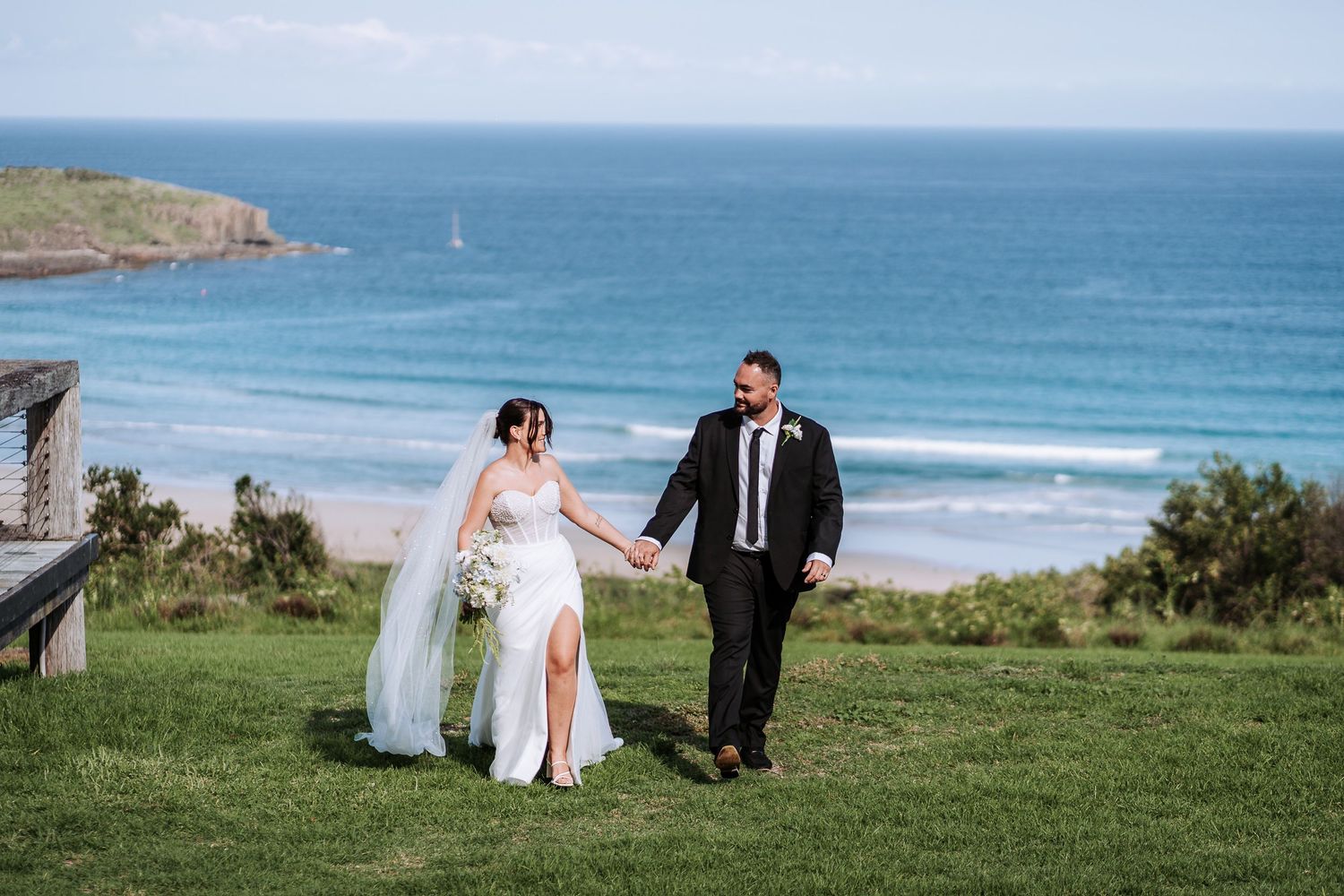 A couple in wedding attire sits together on a grassy cliff overlooking a scenic ocean coastline.
