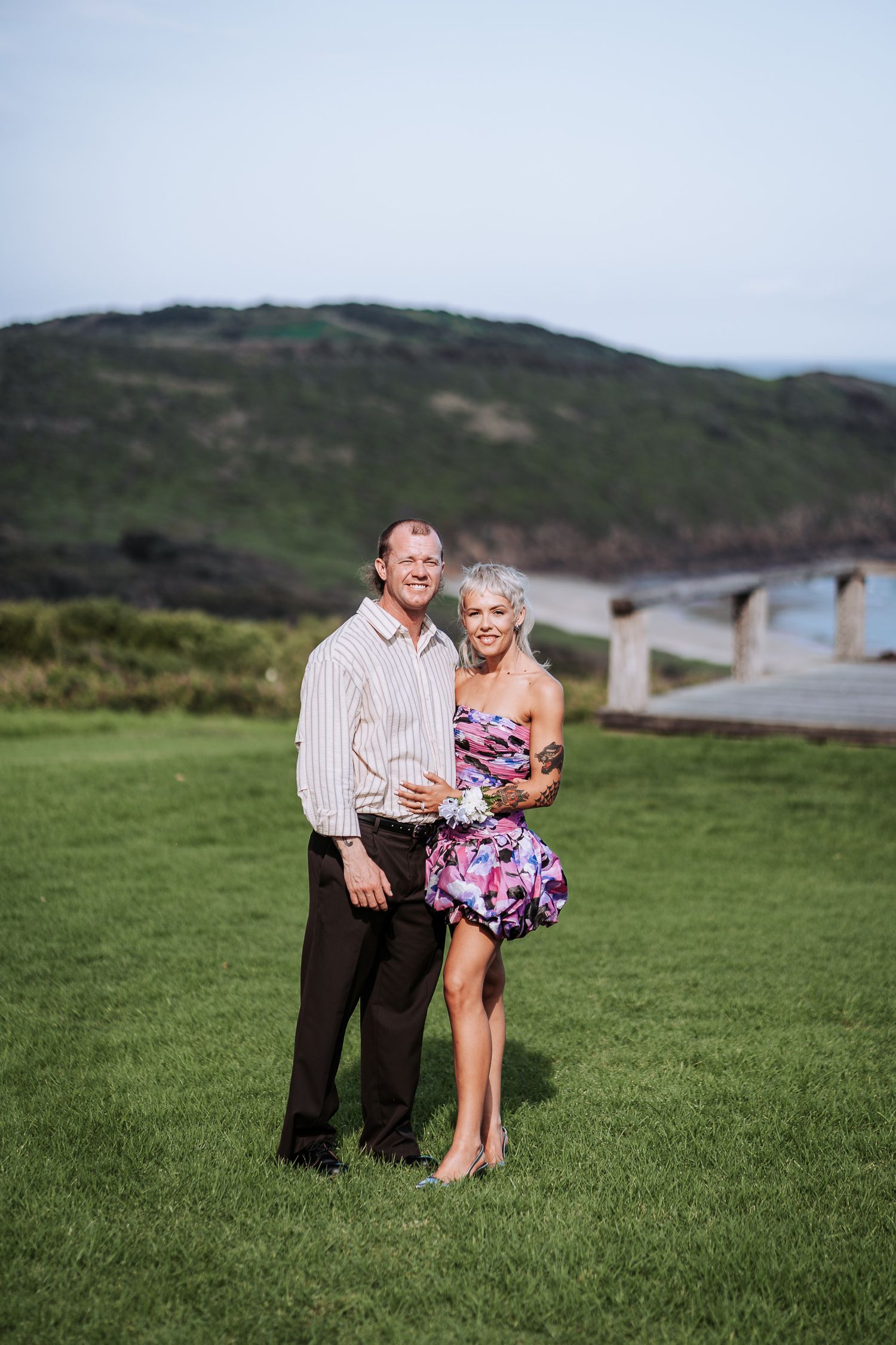 A couple sits together on a grassy hillside overlooking a coastal landscape during golden hour.