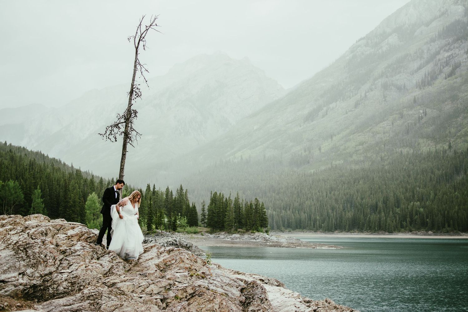 A couple stands on a scenic mountain overlook foggy rocky cliffs and a turquoise lake in Banff  Canadian Rockies. At Lake Minniwanka
