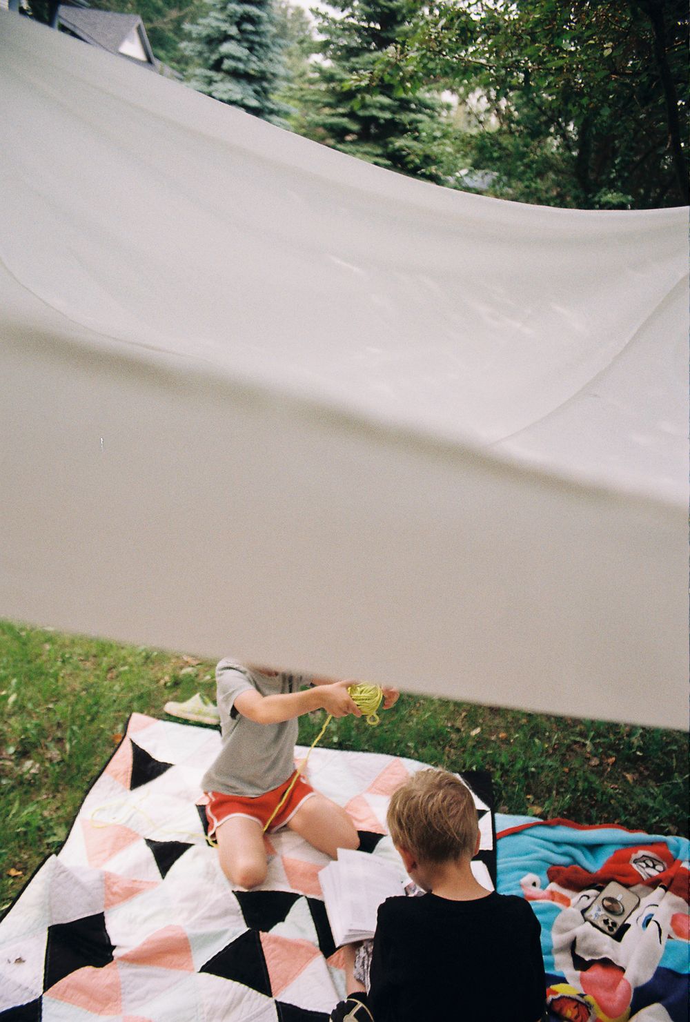 Two children playing under a white sheet canopy on a colorful quilt blanket in a backyard.