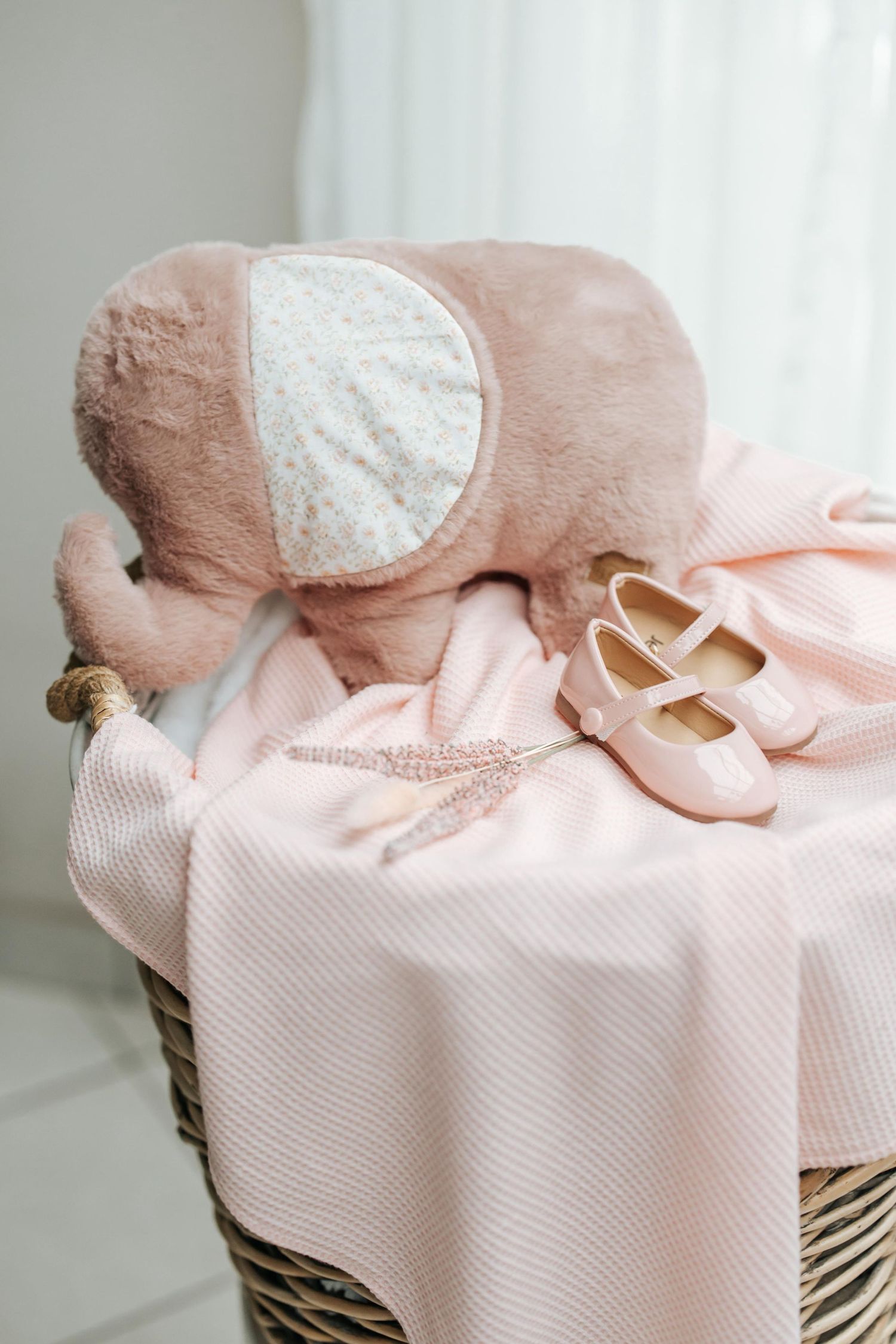 Pink and white baby shoes rest on a striped blanket with a plush teddy bear accessory in soft pastel tones.