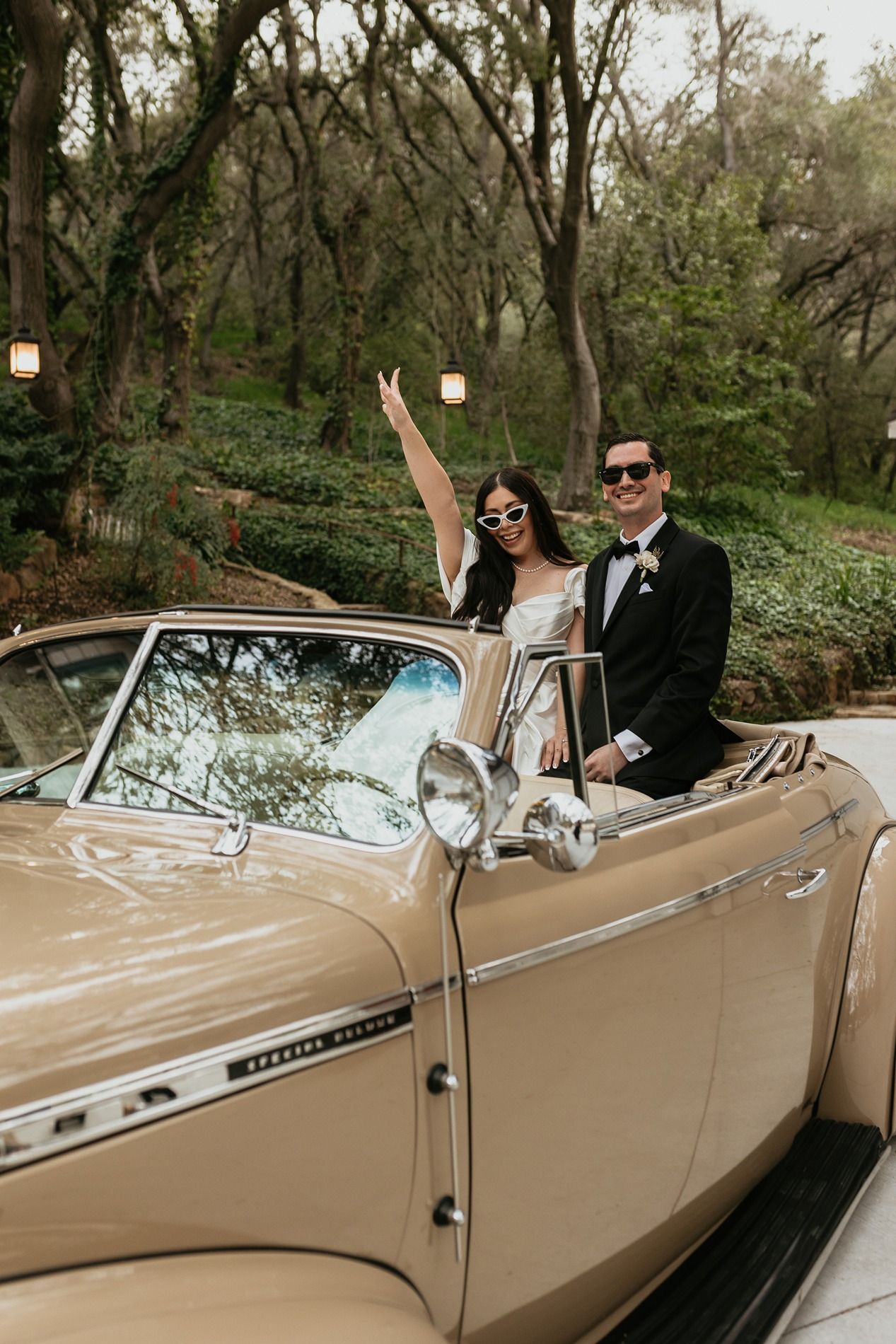 A classic champagne-colored convertible car parked in a wooded area during a formal event at The Oaks at Duncan Lane.