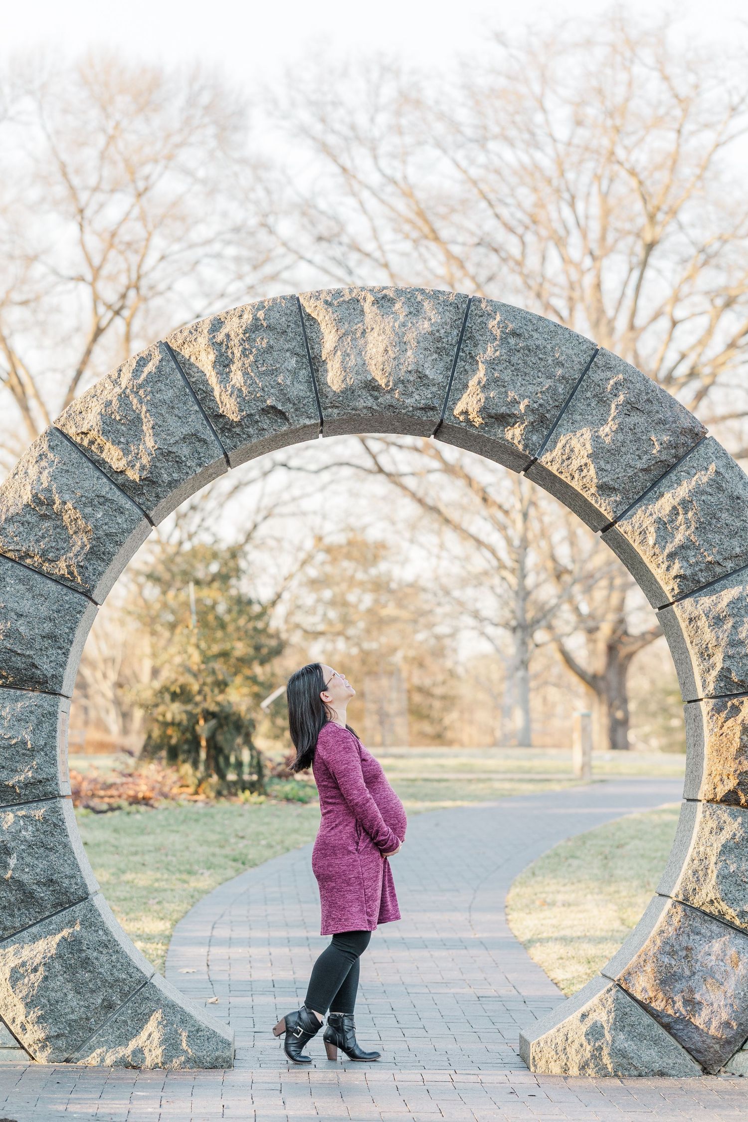 A person in a pink coat walks through a curved stone archway in a park during winter.