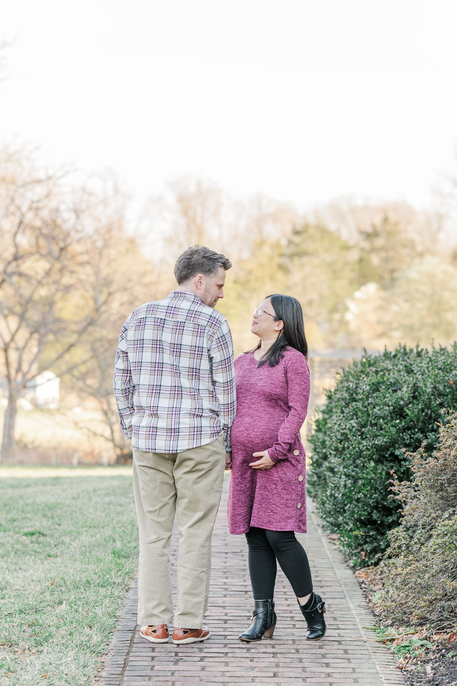 A couple walks together on a brick path surrounded by green shrubs and autumn foliage in a park setting.