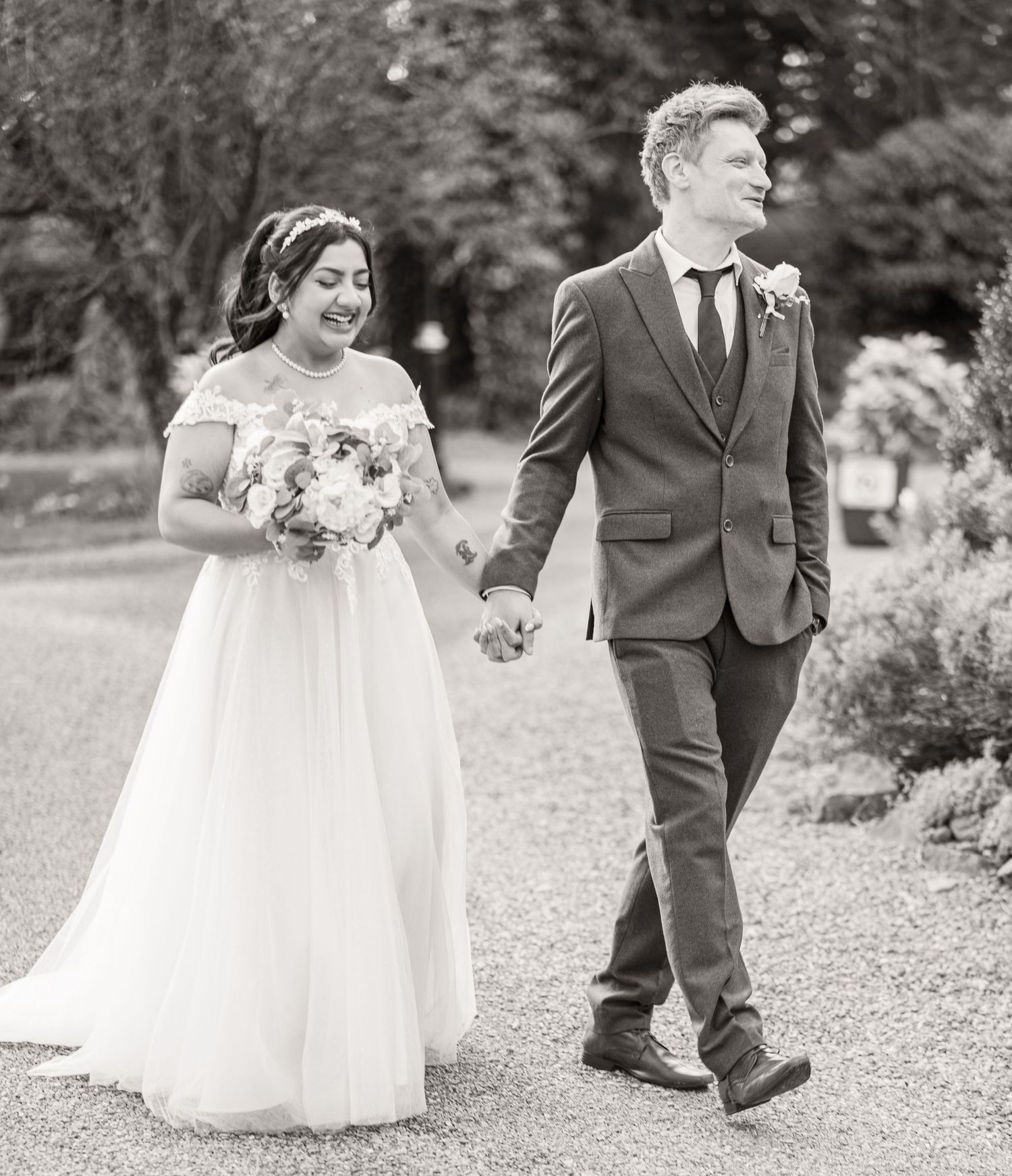 A couple in wedding attire walks along a garden path in black and white photography.