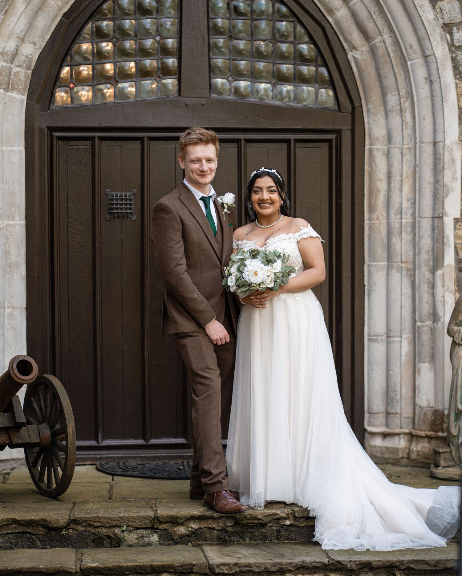 A wedding couple and wedding party pose in front of an elegant stone archway entrance with gothic architecture details.