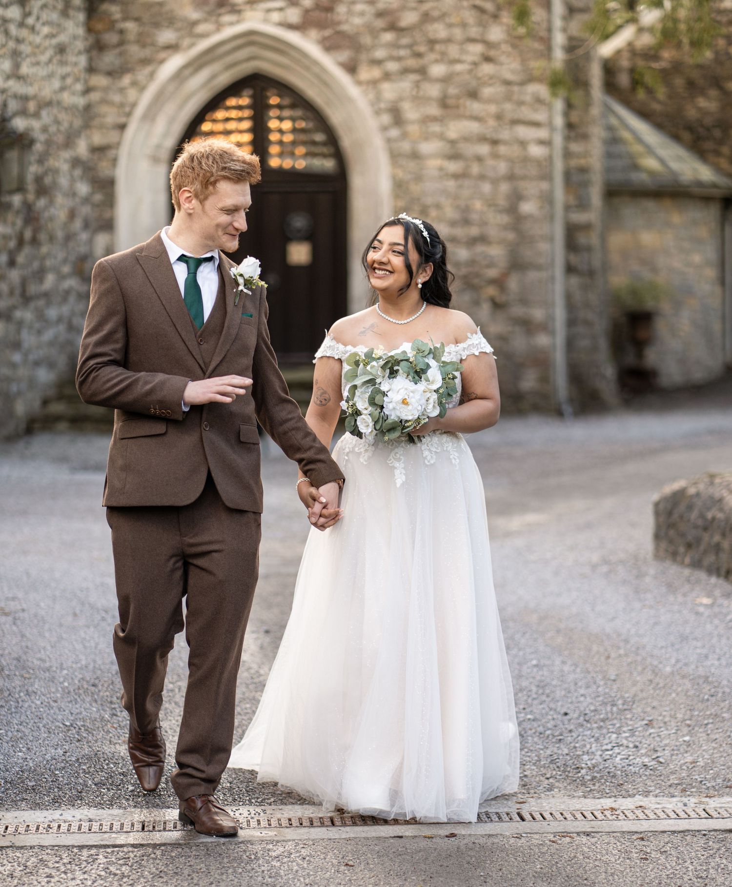 A couple in wedding attire share a tender moment on a stone path outside a historic church building.