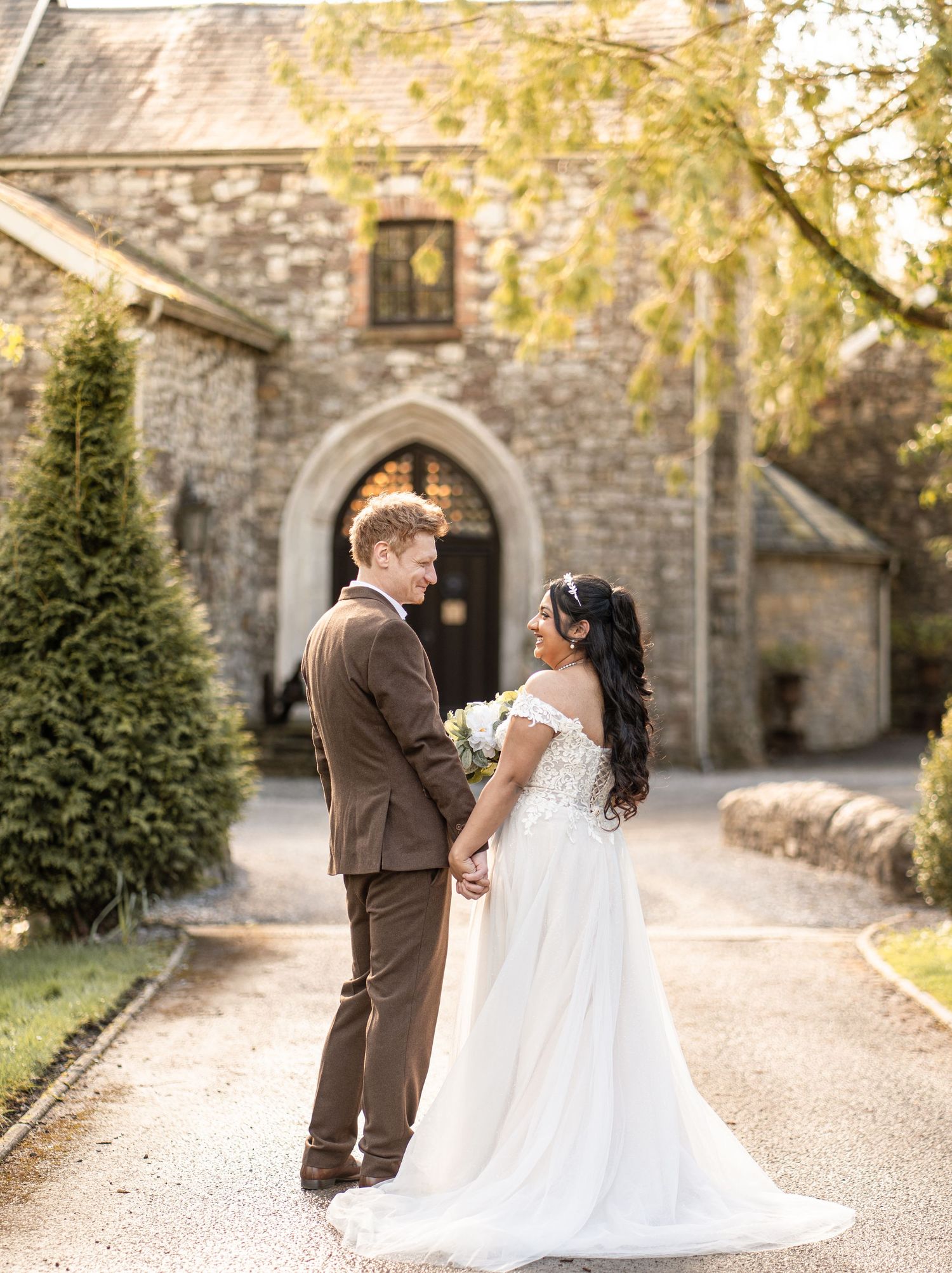 A couple in brown and white wedding attire poses together on an autumn path with stone walls and evergreens.