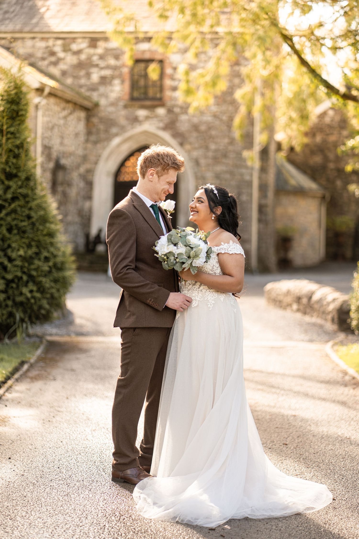 A joyful wedding couple shares an intimate moment outdoors with stone architecture and autumn foliage in the background.
