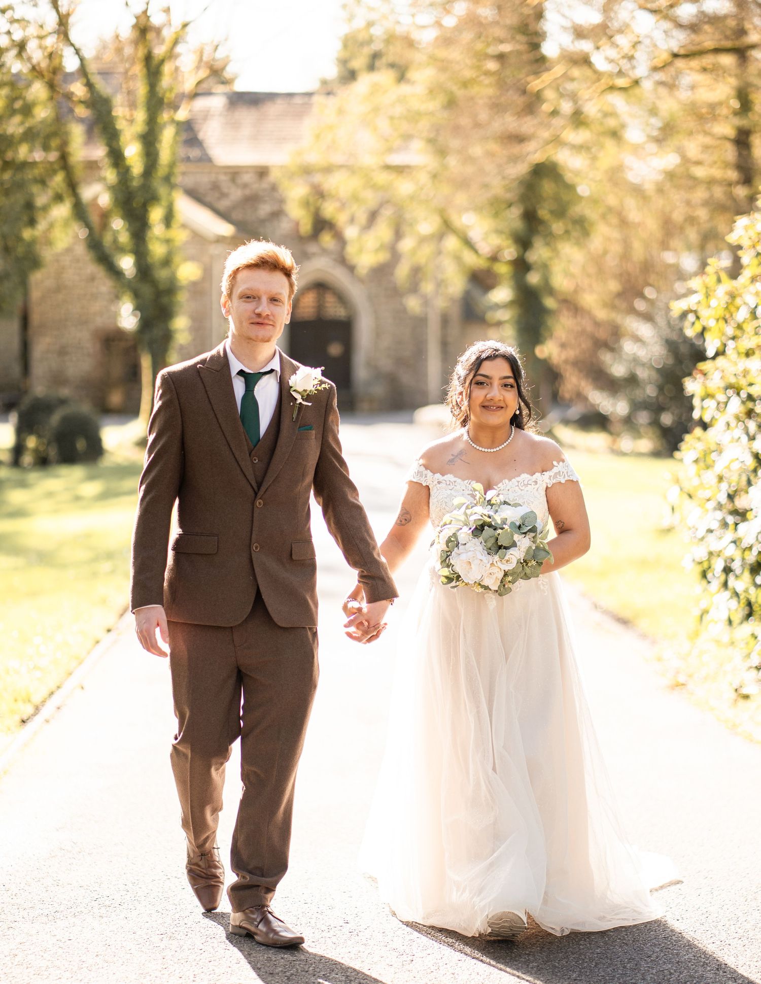 A couple in wedding attire walks together on a sunlit path through autumn trees.