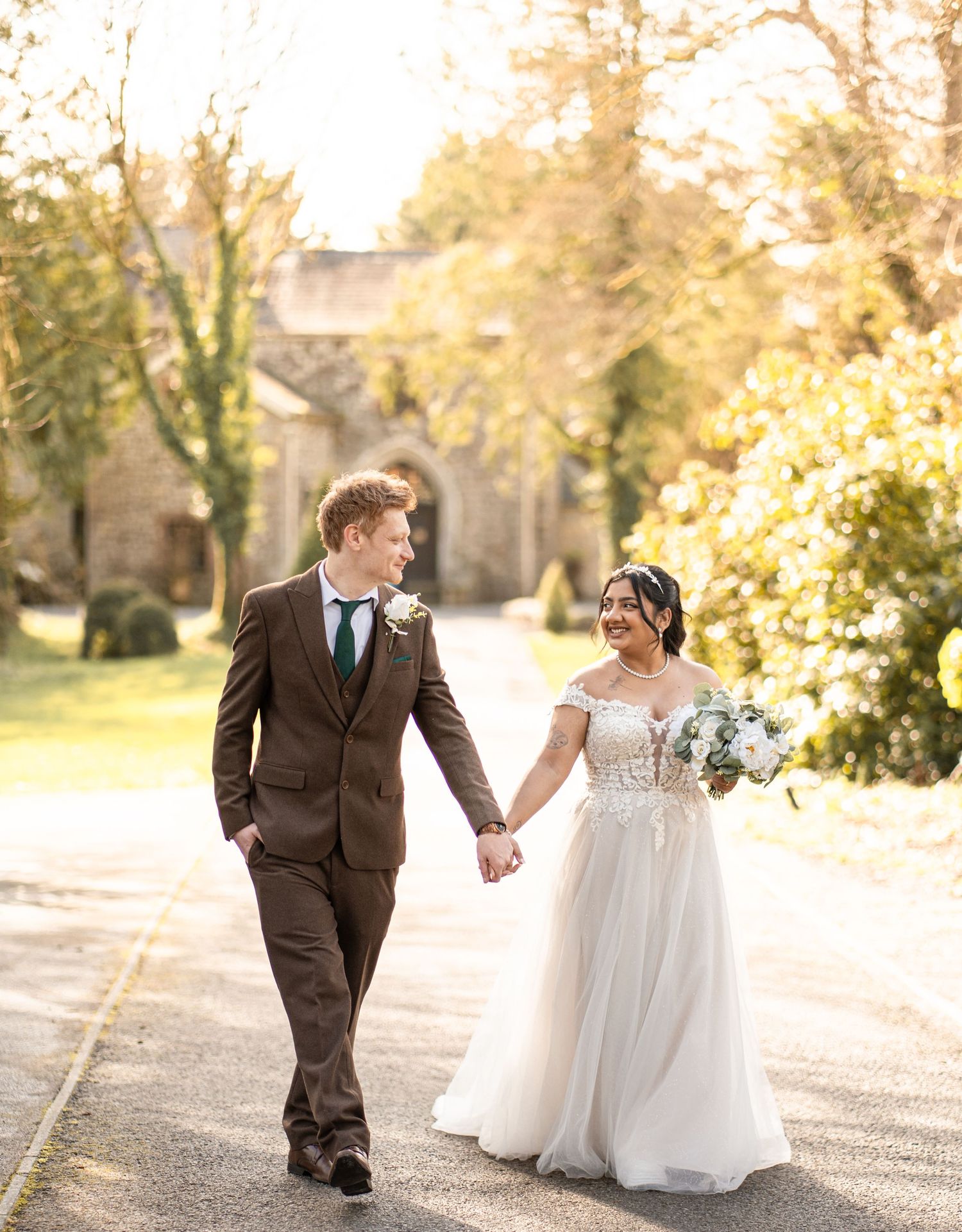 A bride and groom share a romantic moment while walking together on a sunlit autumn path.