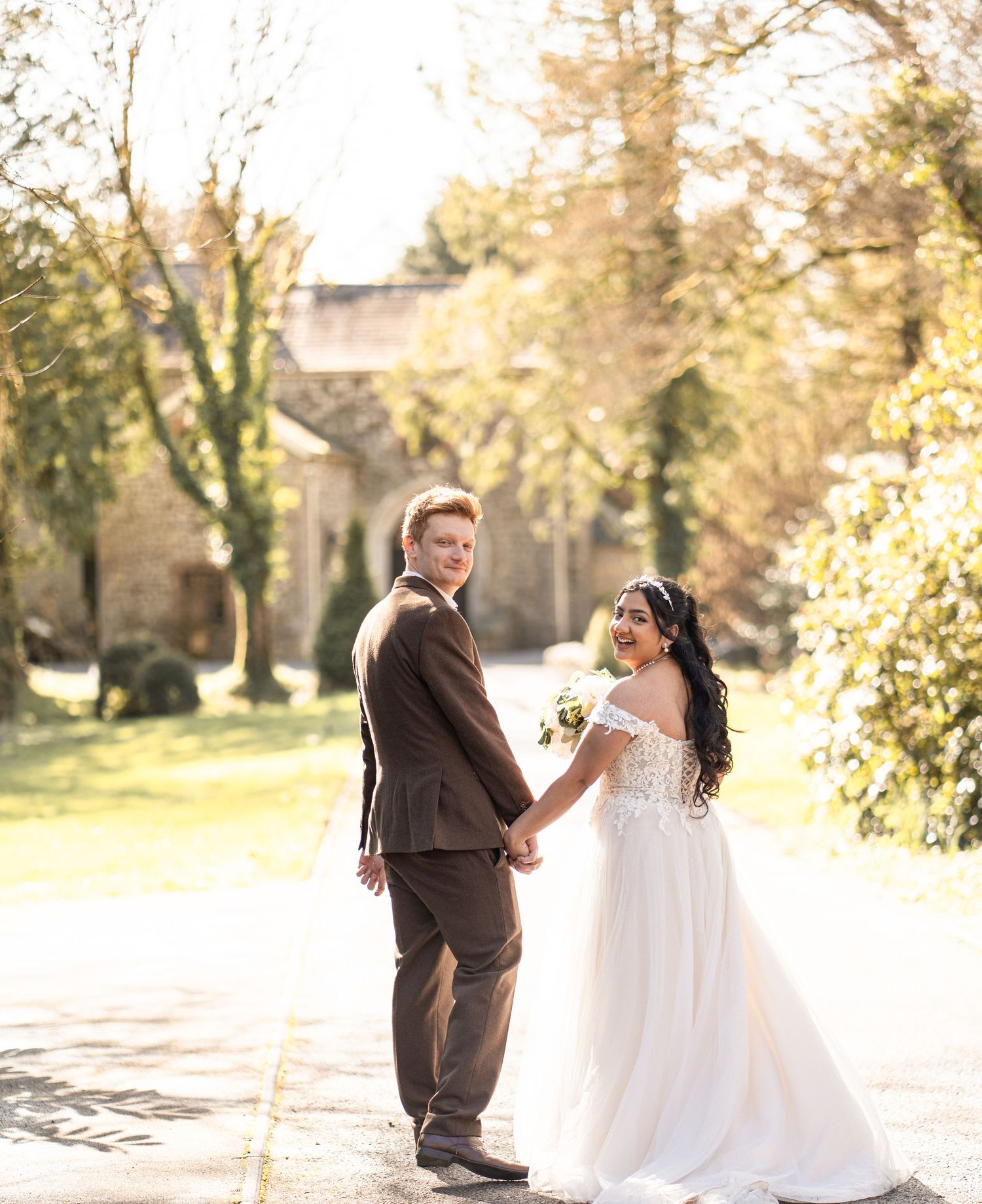 A couple in wedding attire holds hands while standing on a sunlit path surrounded by autumn trees and foliage.