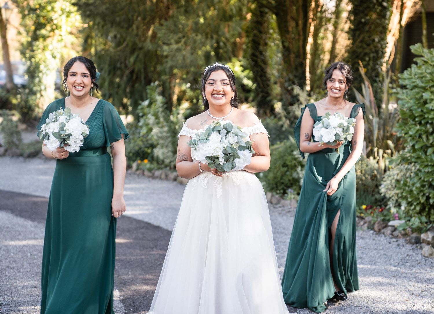 Wedding party in emerald green dresses and white gown pose together in a wooded outdoor setting.