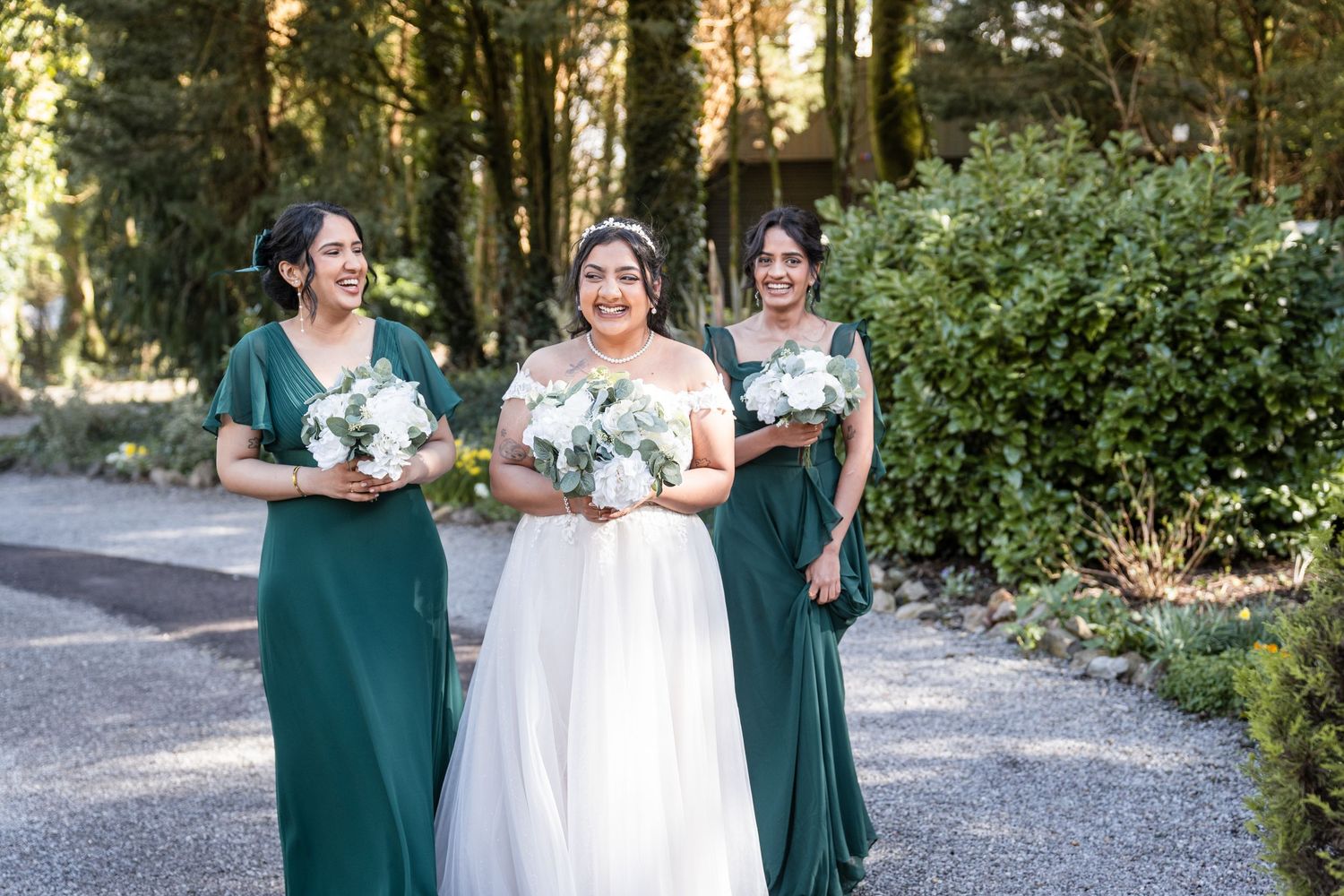 Wedding party in emerald green dresses and white gown pose together in outdoor garden setting.