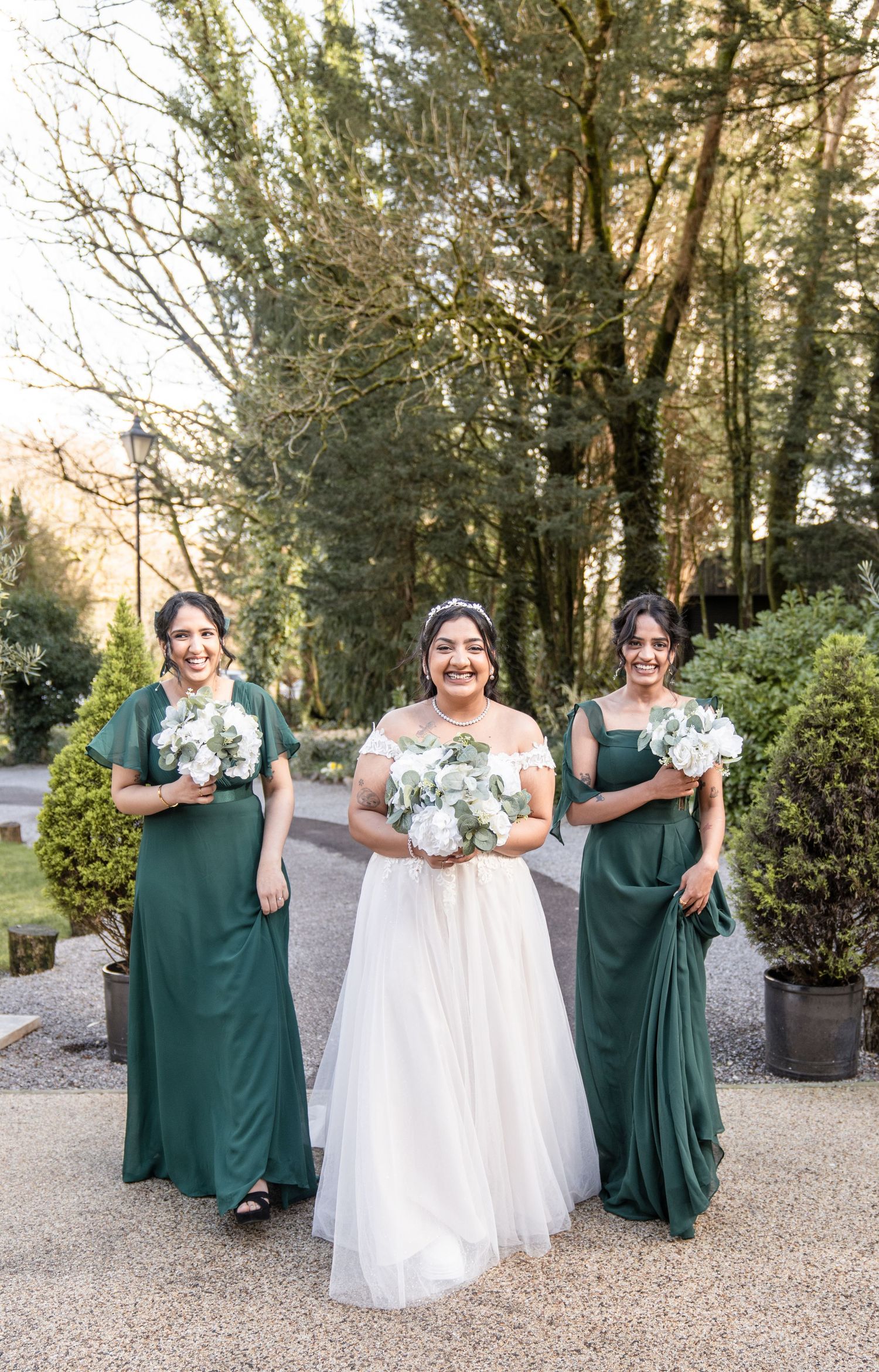Wedding party in emerald green dresses and white gown pose together on gravel path surrounded by trees.