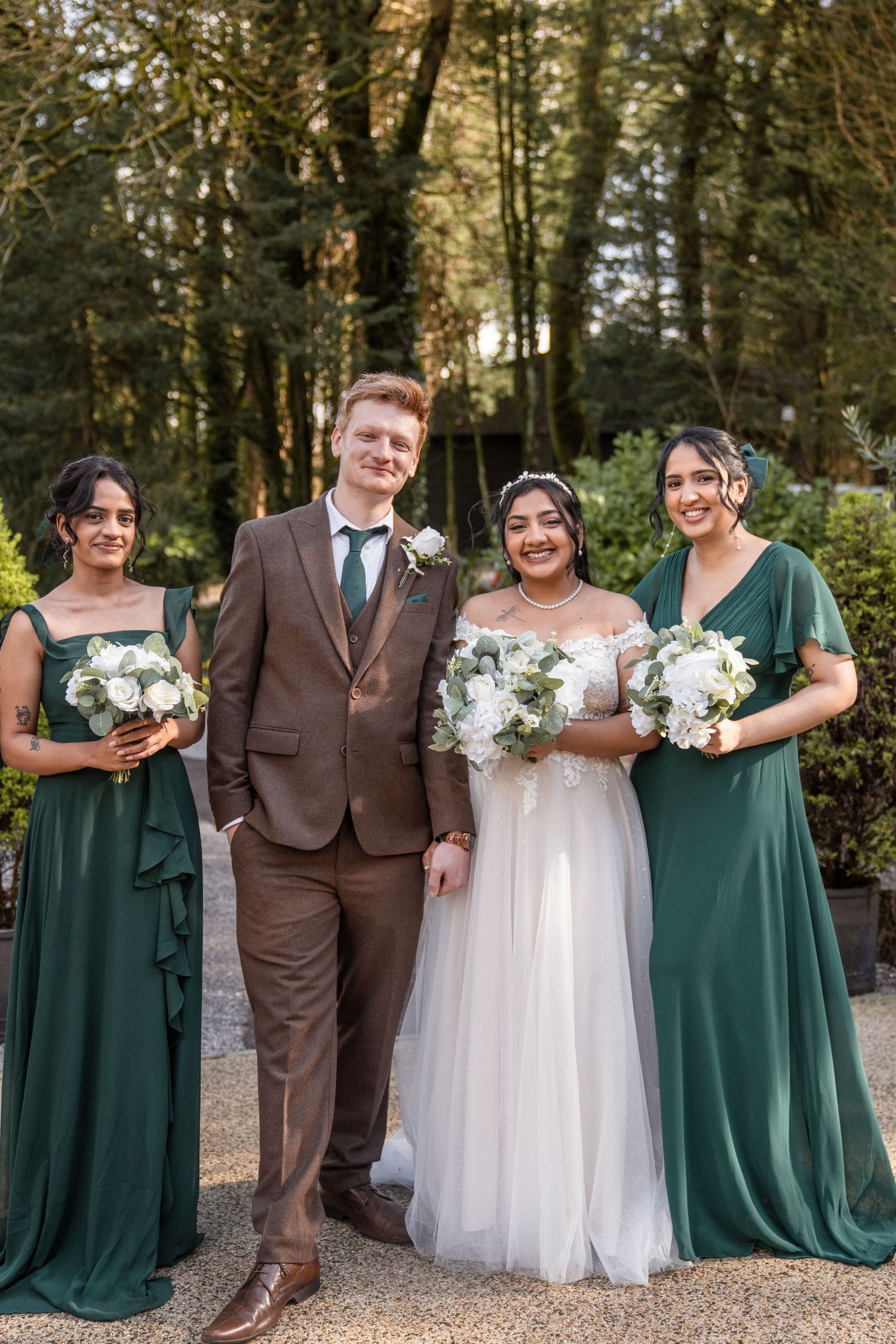 Wedding party poses in a wooded setting with emerald green bridesmaid dresses and brown suit against natural backdrop.