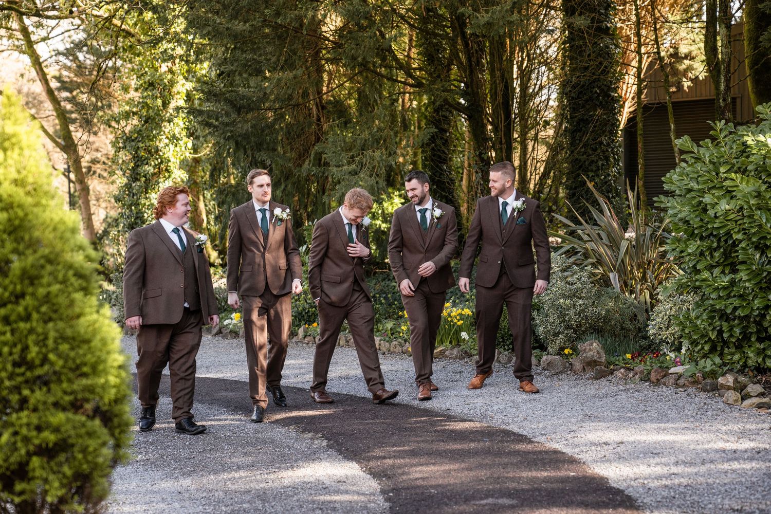 A wedding party walks through a wooded path in brown suits on a sunny afternoon.