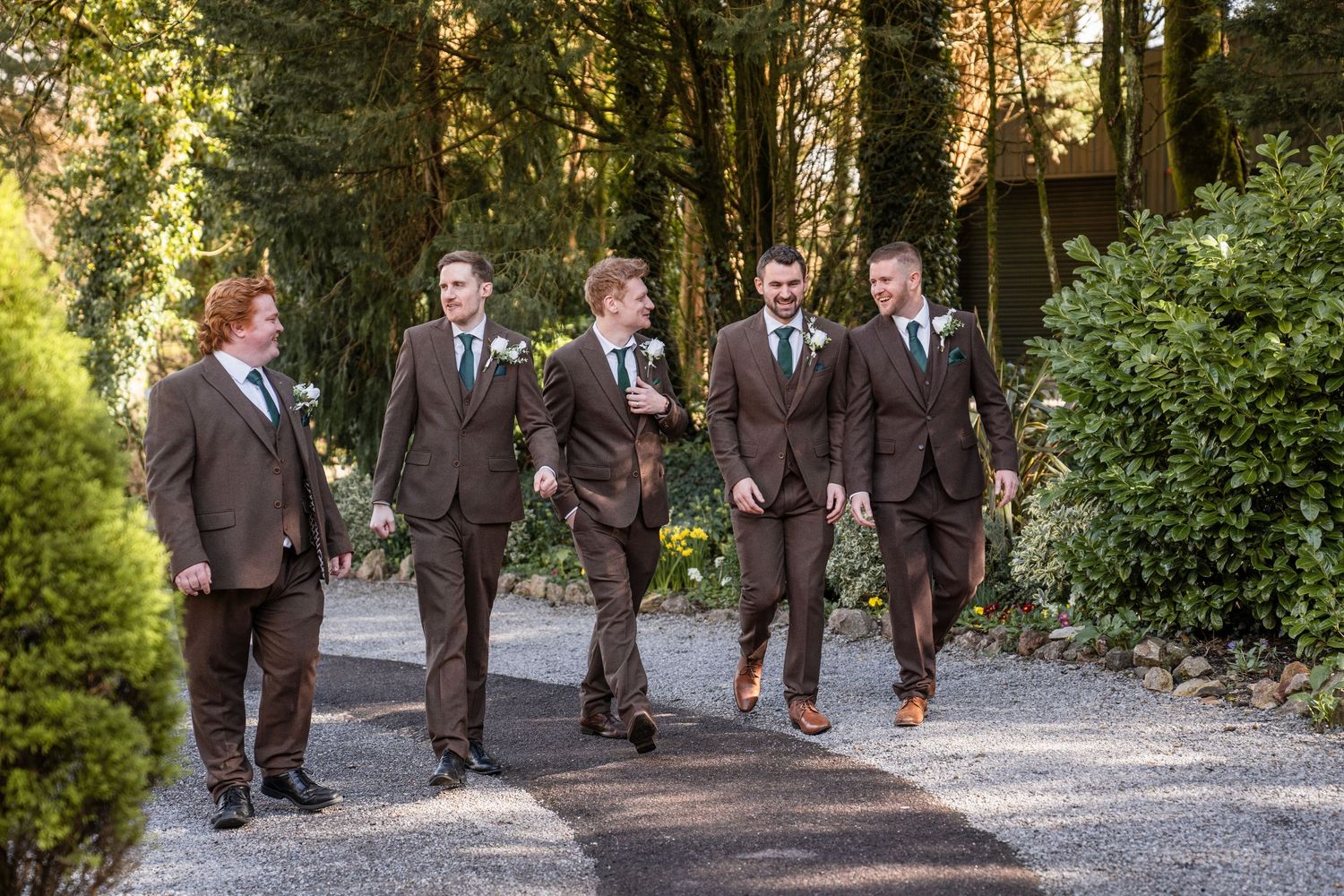 A group of men in matching brown suits walk together on a tree-lined path during a wedding celebration.