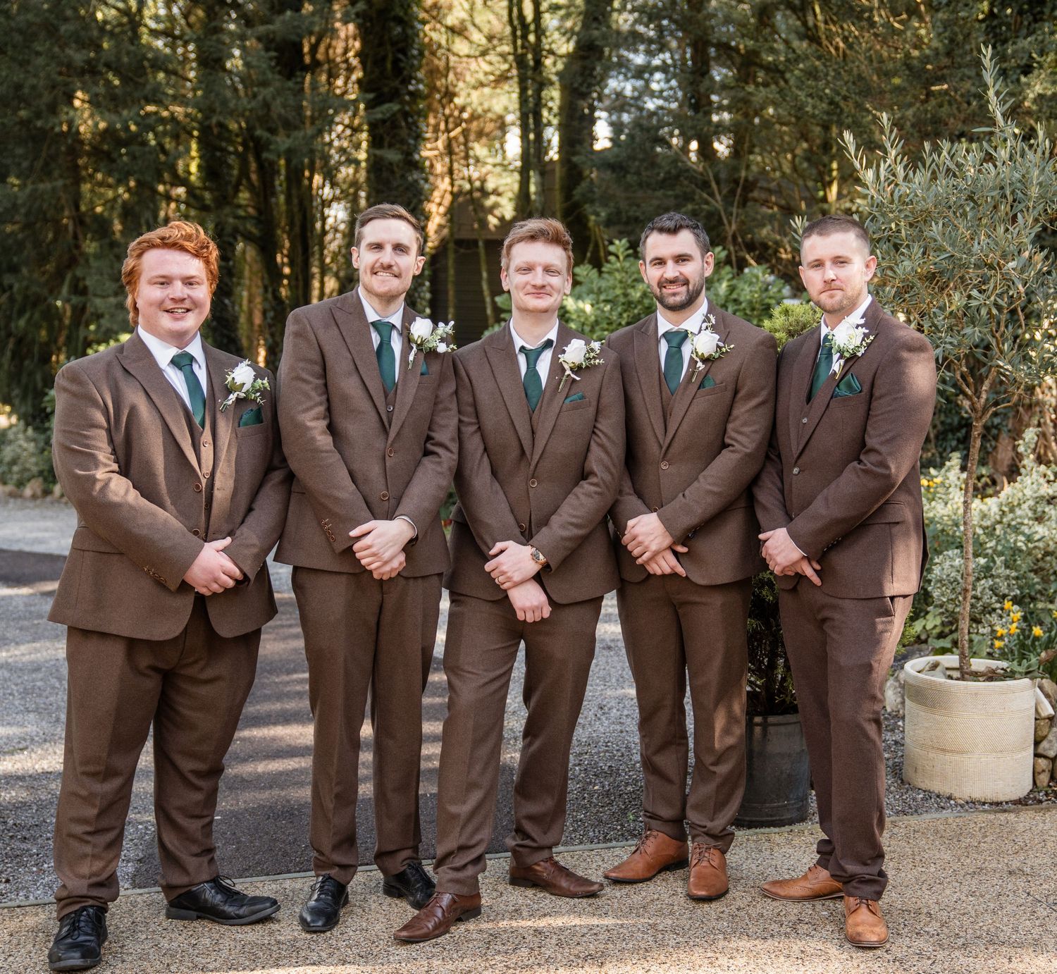 A group of five groomsmen pose together in matching brown suits and boutonnieres at an outdoor wedding venue.