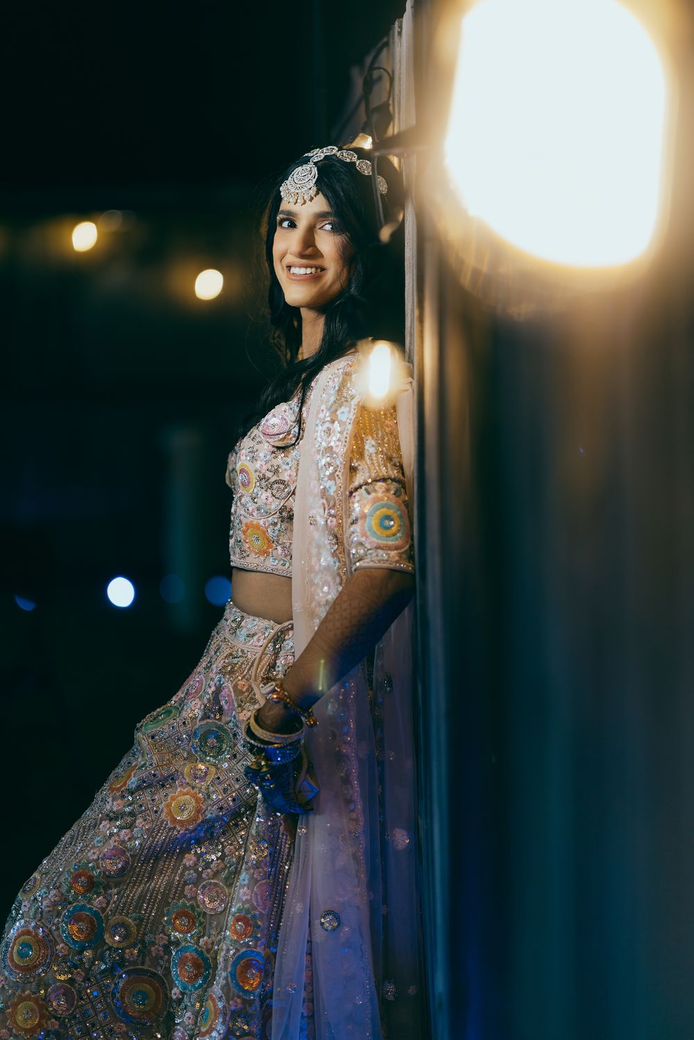 A person wearing an ornate Indian lehenga with intricate embroidery poses in a dimly lit setting with glowing lights.
