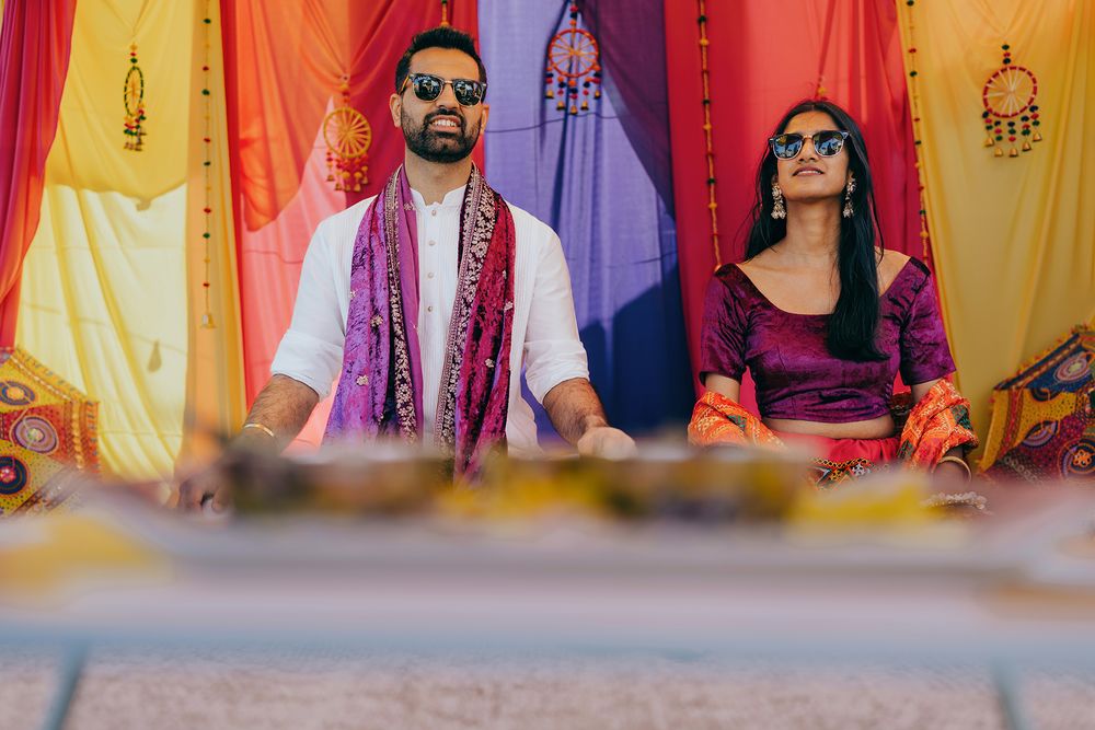 Couple in traditional Indian wedding attire sits together against vibrant yellow, orange and purple backdrop during ceremony.