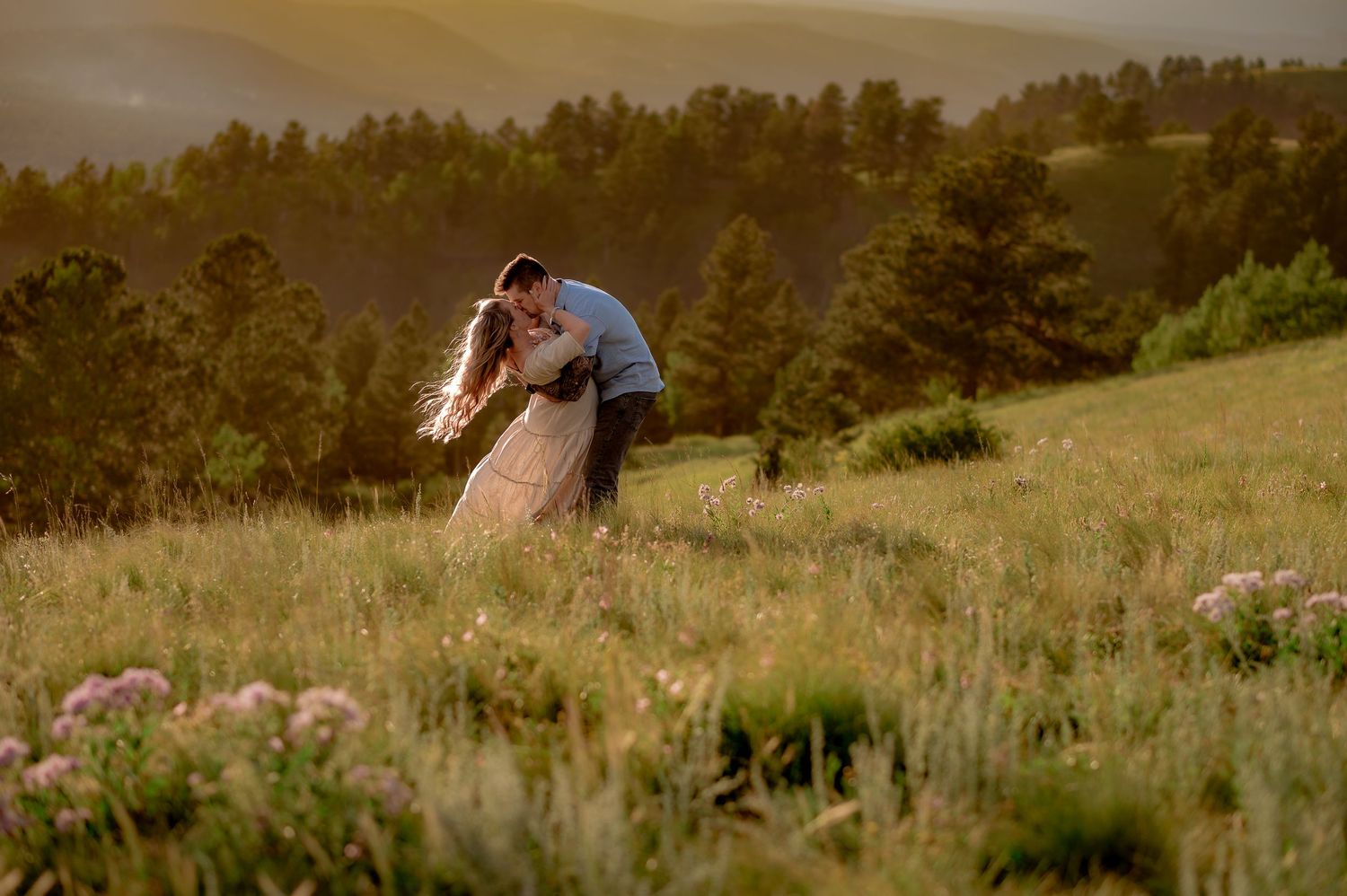 A couple shares a romantic moment in a scenic meadow at sunset with mountains in the background.