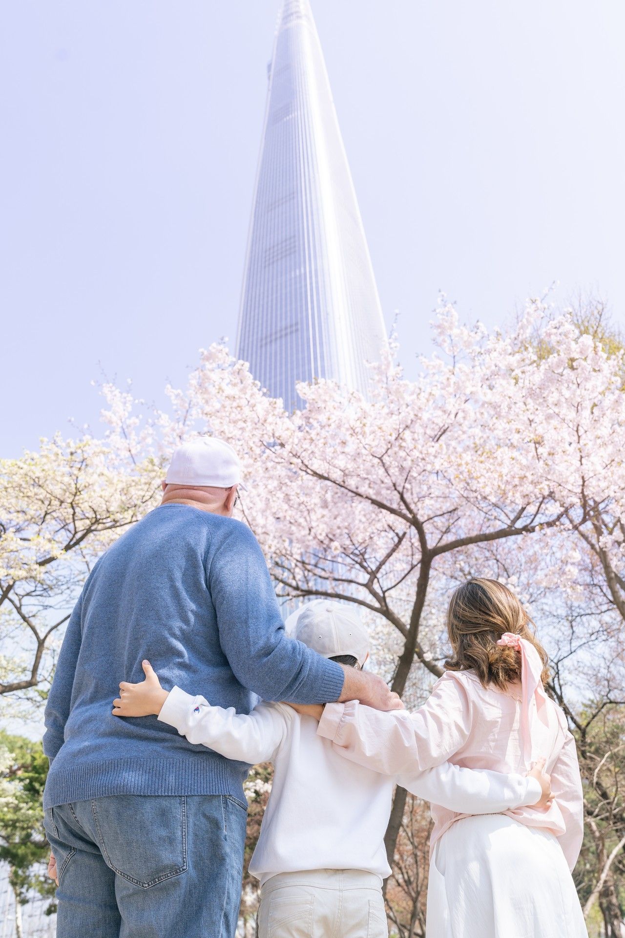 Three people walk together arm in arm under blooming cherry blossom trees with a tall tower visible in the background.