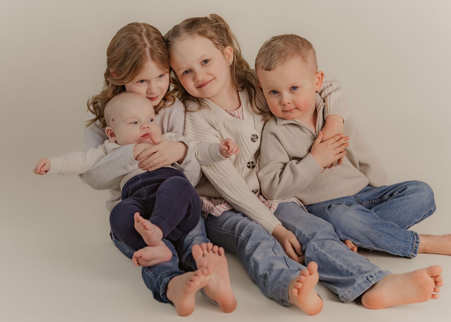 Four children in coordinating beige sweaters and jeans sit together on a white backdrop showing bare feet.