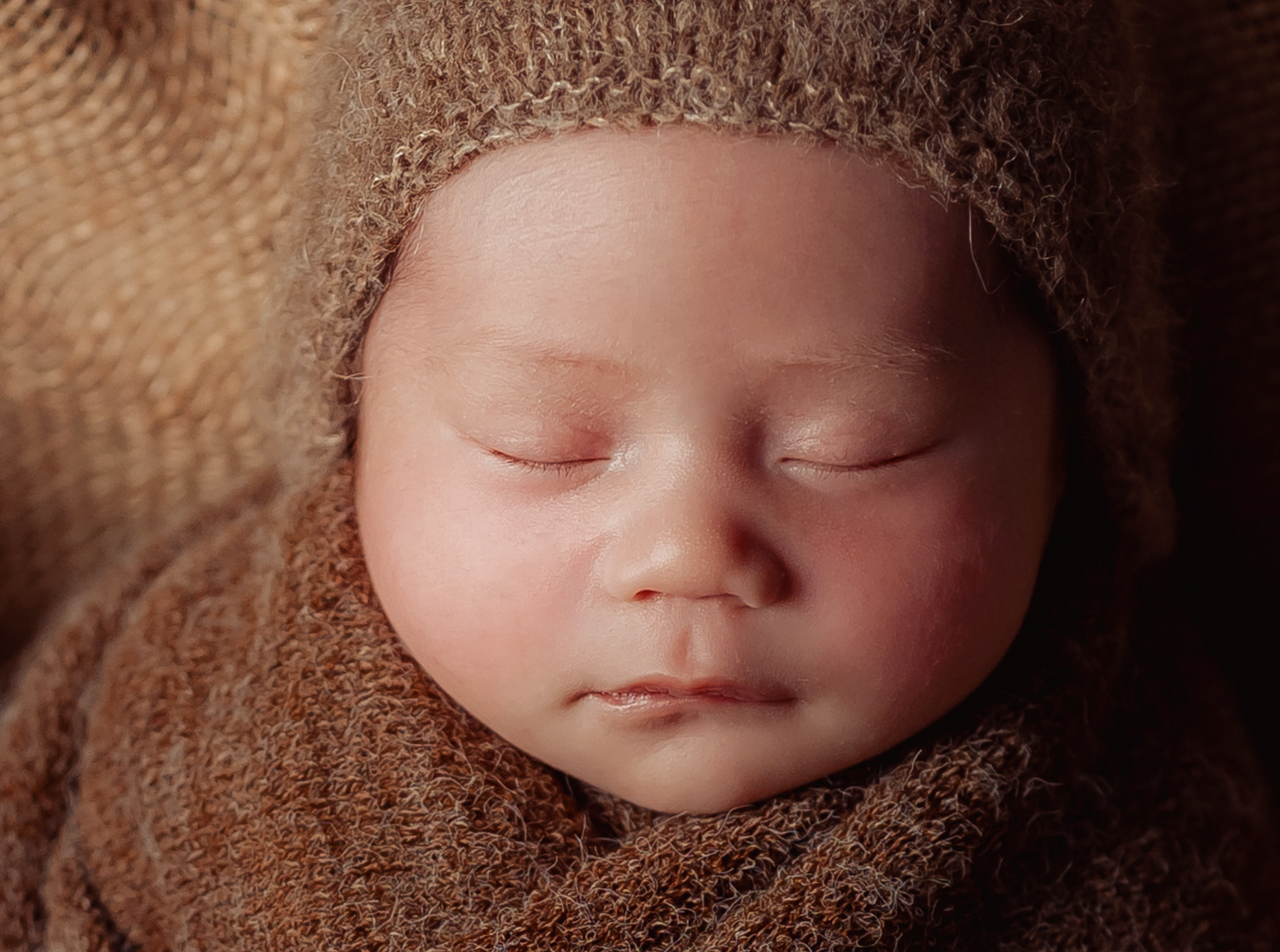 Close-up portrait of a sleeping newborn wrapped in soft brown fabric with delicate lighting.