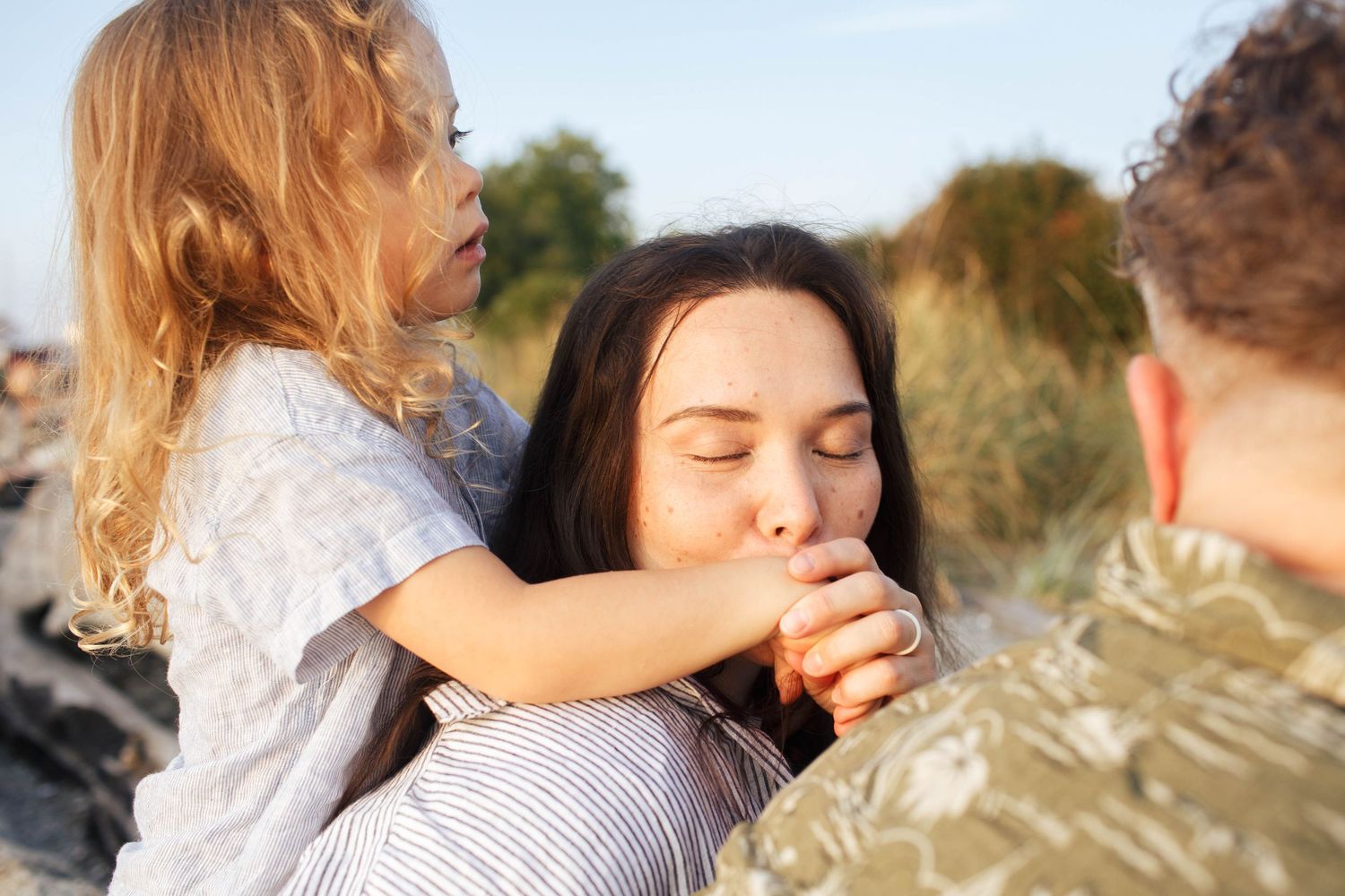 Close-knit family photographer in Seattle WA area: A mother kisses her sons hand during a family photo session at the beach in the summer