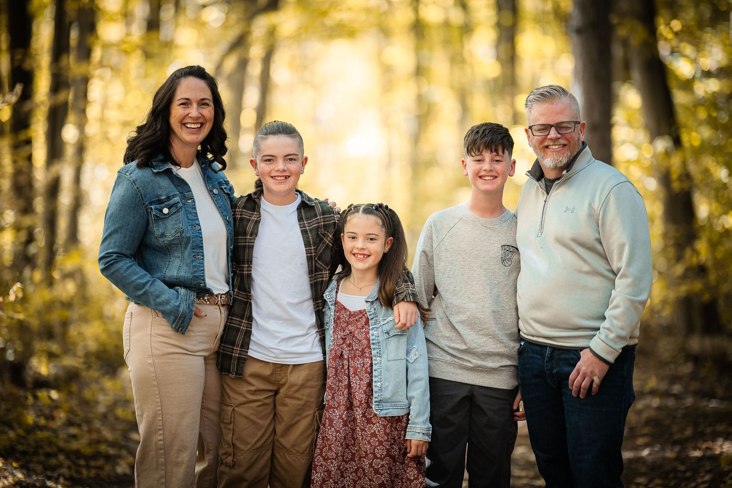 A family group poses together outdoors in a sunlit autumn forest setting wearing coordinated casual attire.