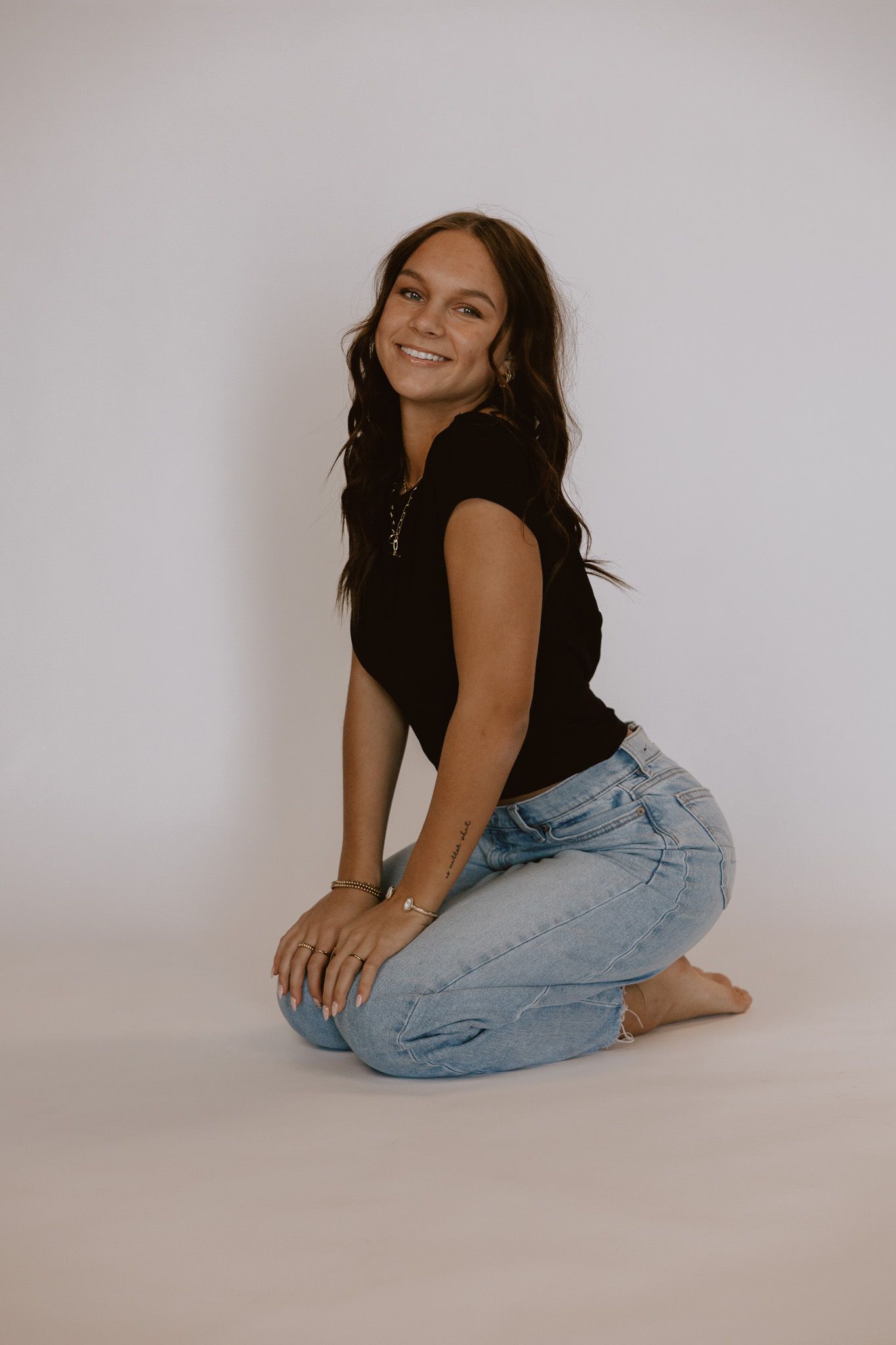 Smiling young woman in black top and light blue jeans kneeling on white studio background.