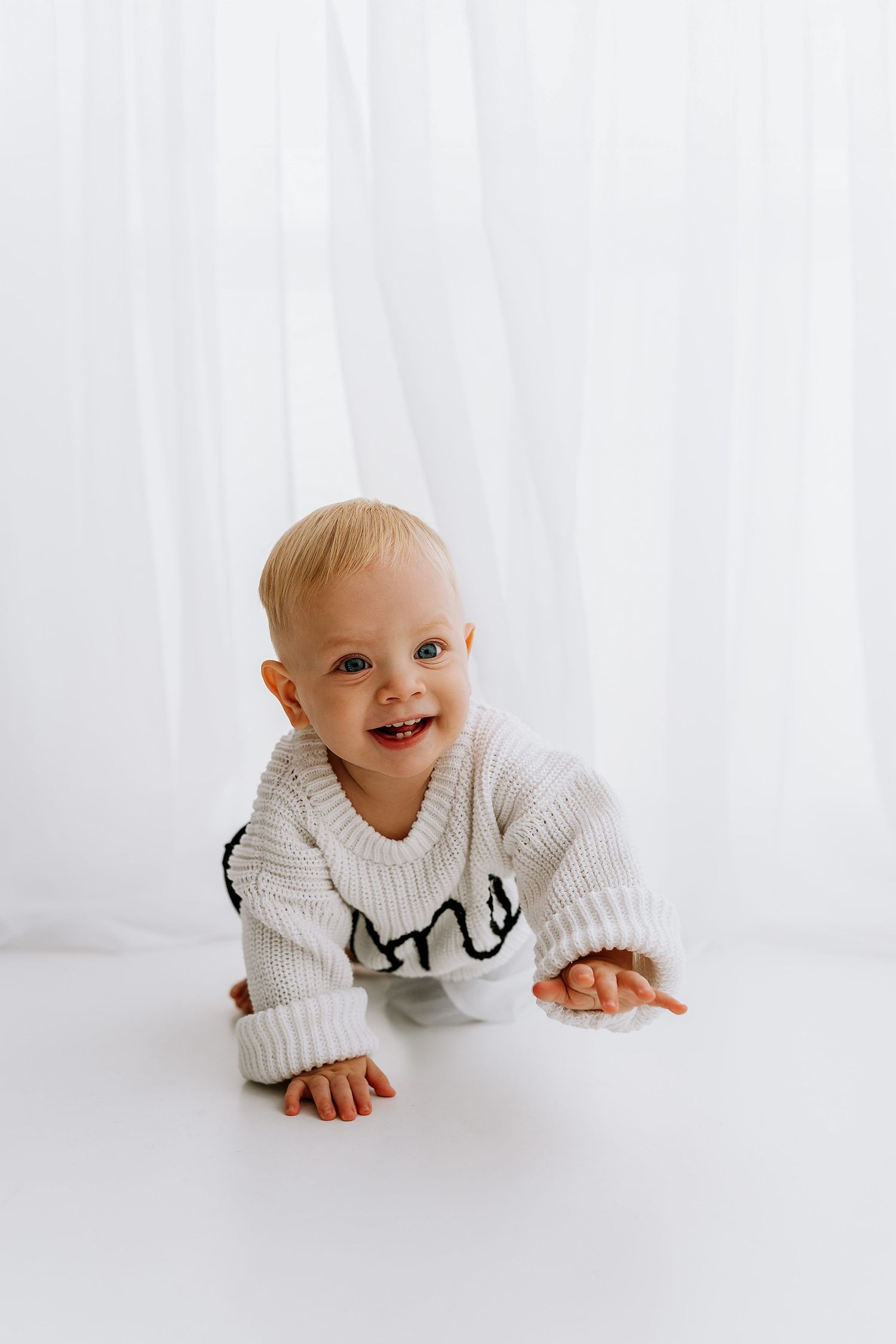 Baby in white knit sweater crawls and smiles against white backdrop in classic minimalist studio photo.