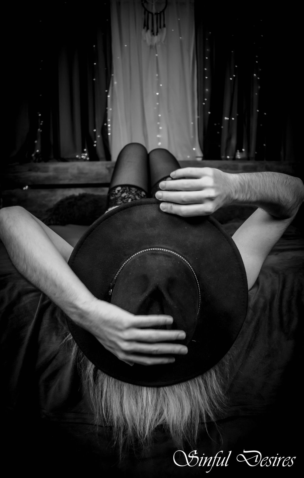 Black and white photo showing a person laying down wearing a dark hat with fairy lights and dreamcatcher in background.