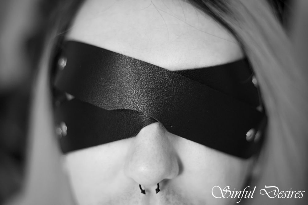 Close up black and white photo showing someone wearing a black blindfold with metal studs against pale skin.