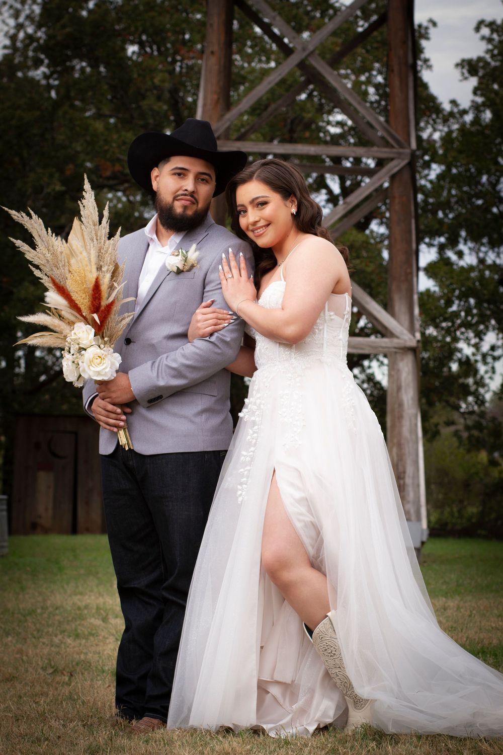 Wedding couple posing outdoors with dried pampas grass bouquet under wooden pergola.