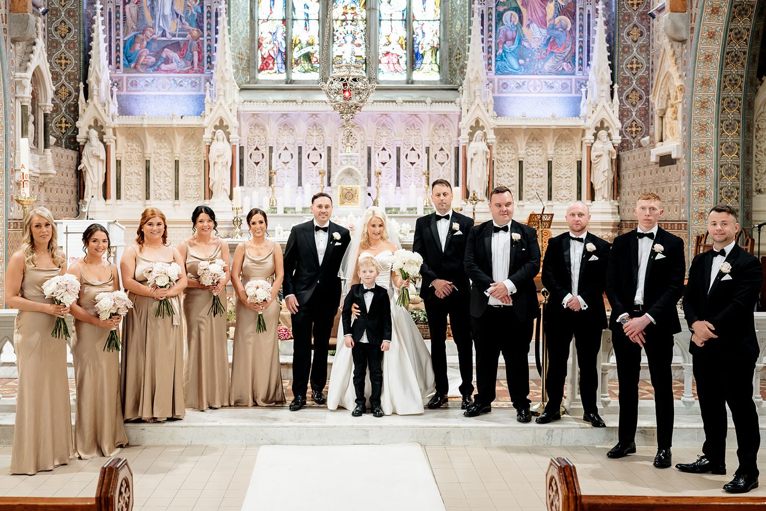 A formal wedding party poses together inside an ornate church with stained glass windows and decorative architecture.