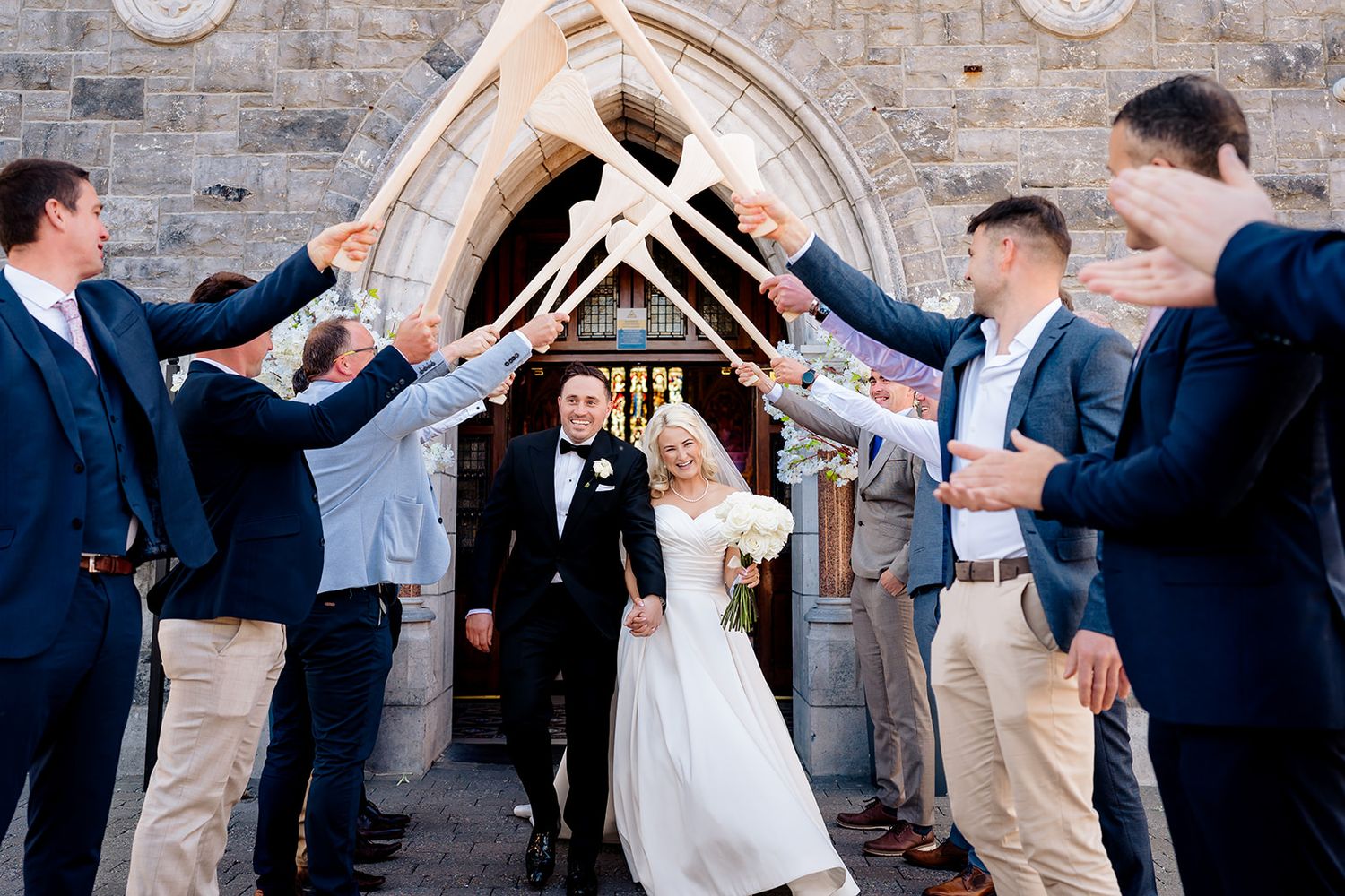 A couple exits a stone church entrance under an archway of raised arms during their wedding ceremony.